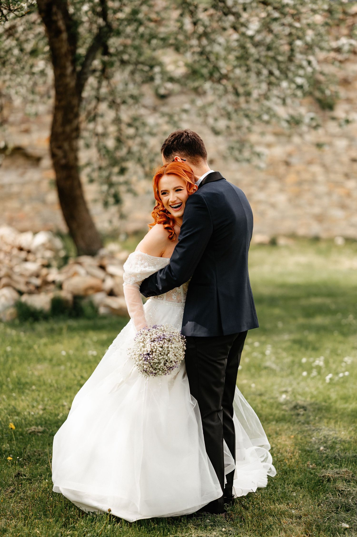 Bride in white off-shoulder gown holding bouquet embraces groom in black suit outdoors near stone wall.