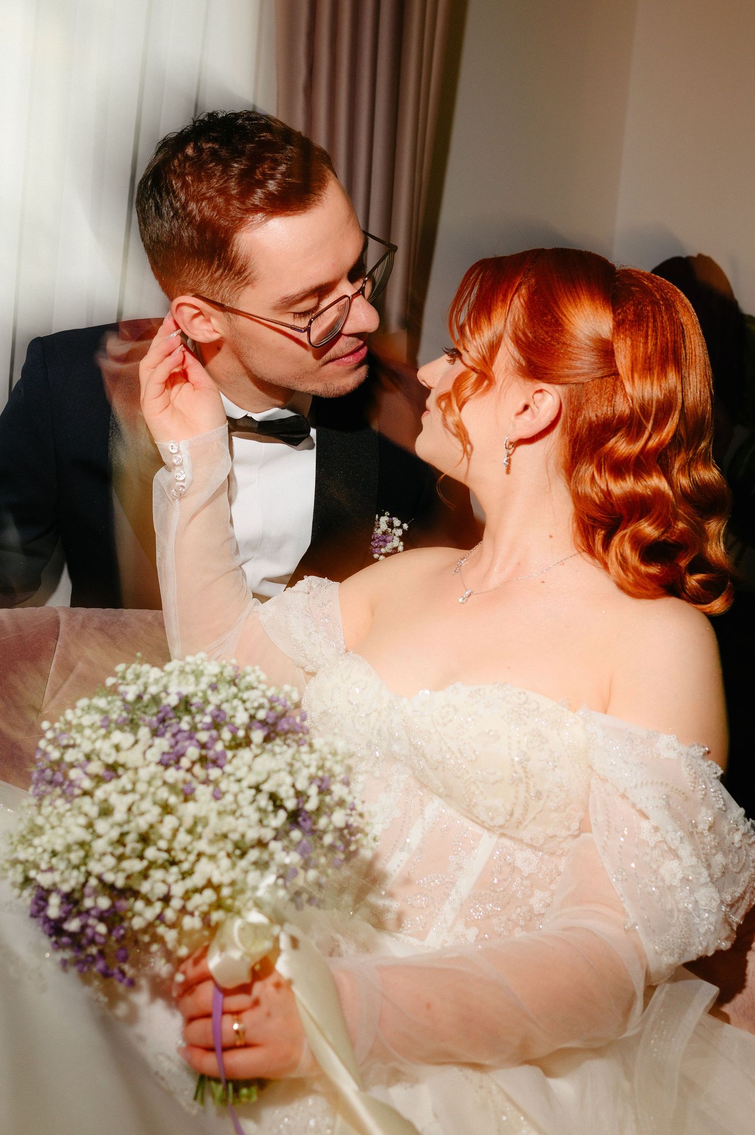 A bride with red hair holding a floral bouquet gazes at her groom in glasses and tuxedo on their wedding day.