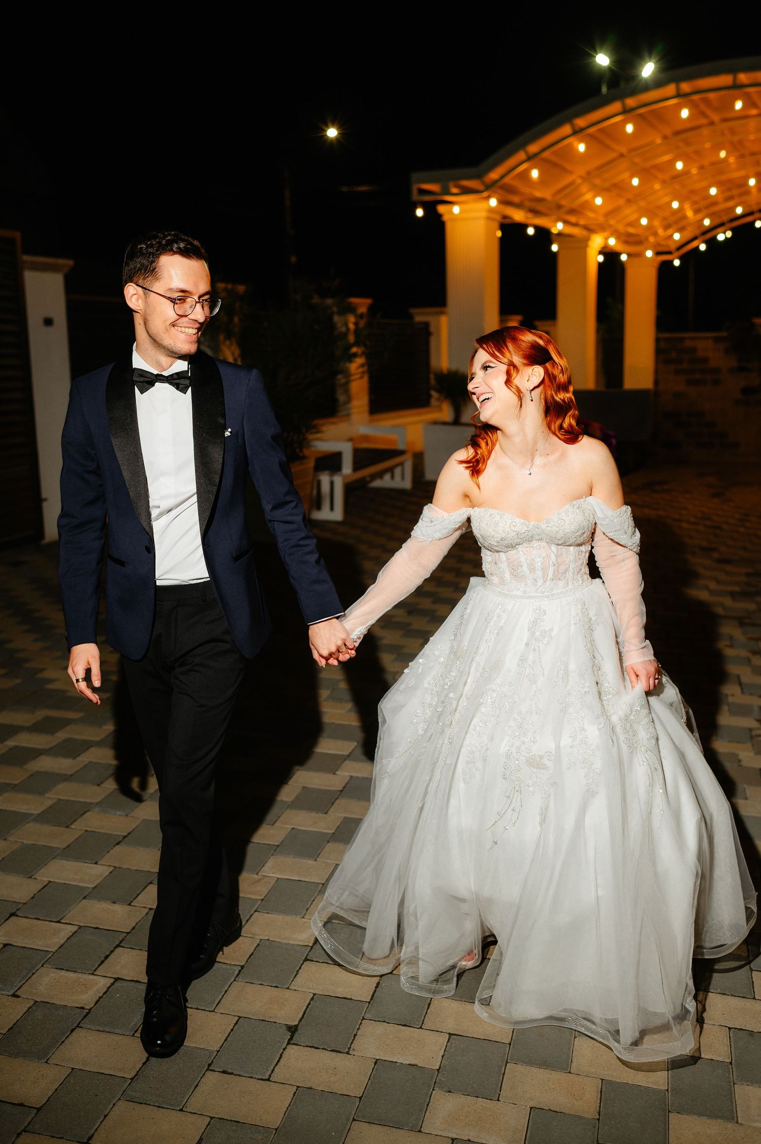 A bride and groom walk hand in hand at night, smiling, she in a white gown, he in a navy tuxedo.