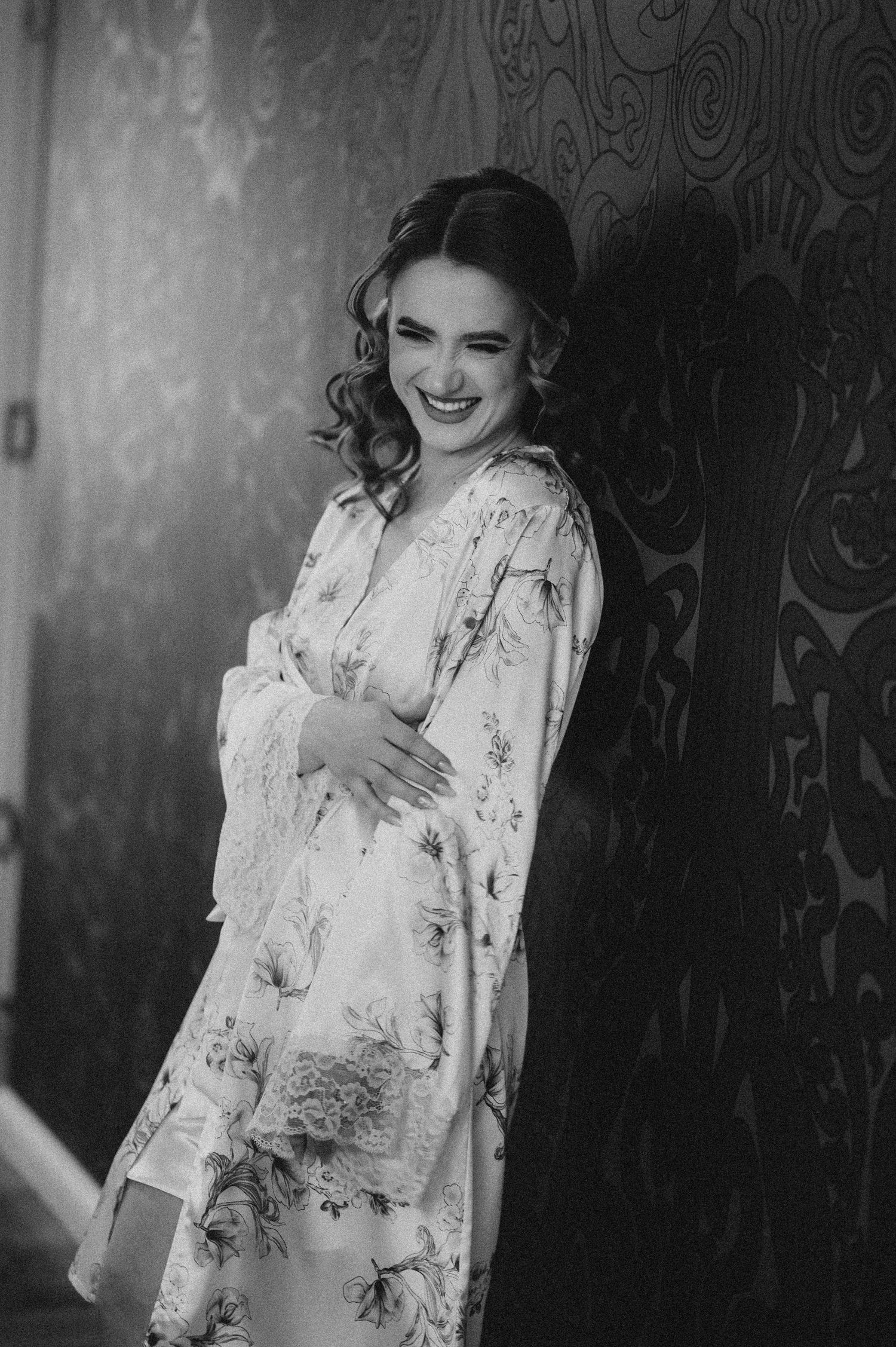 Smiling bride in floral robe leaning against ornate wall in elegant black and white portrait photo.