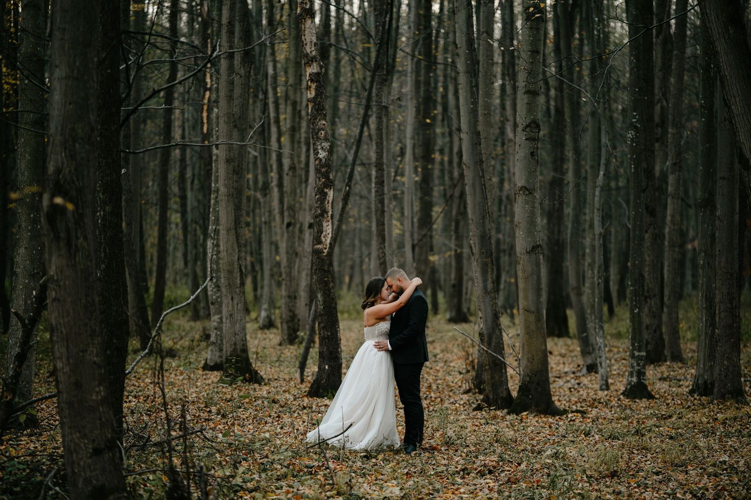 Bride and groom share a romantic embrace in a moody autumn forest, surrounded by tall trees and fallen leaves.