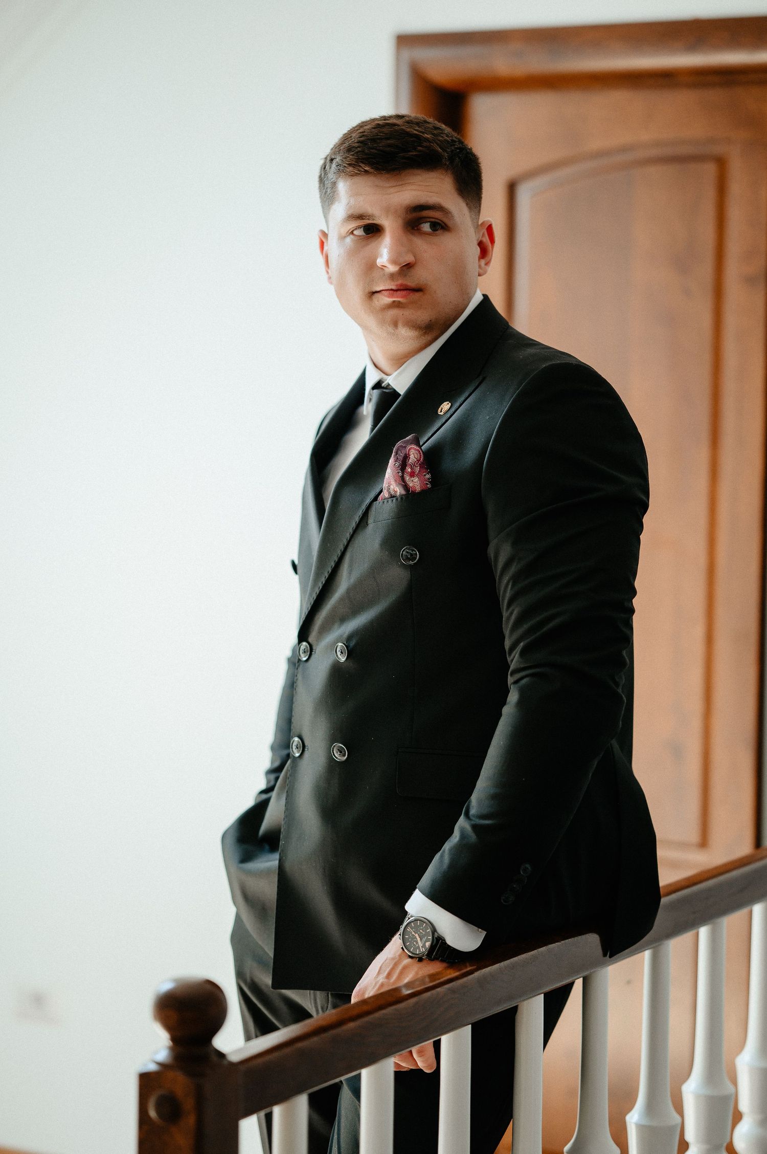 Groom in black double-breasted suit with pink pocket square leaning on staircase railing.