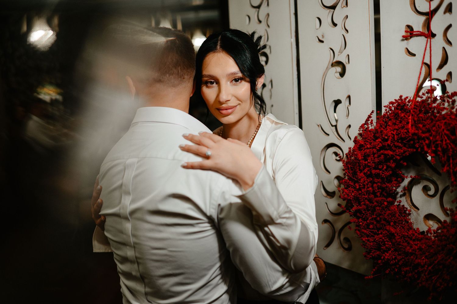 Bride in white satin blouse tenderly embraces groom near ornate white wall with red wreath.