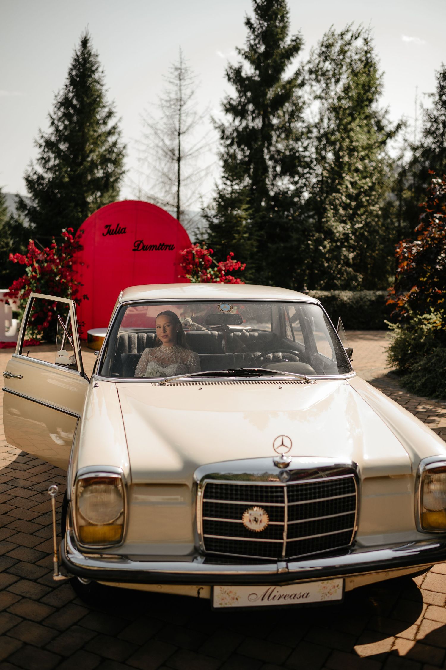 Bride driving classic white vintage Mercedes-Benz wedding car decorated with red floral arrangements.