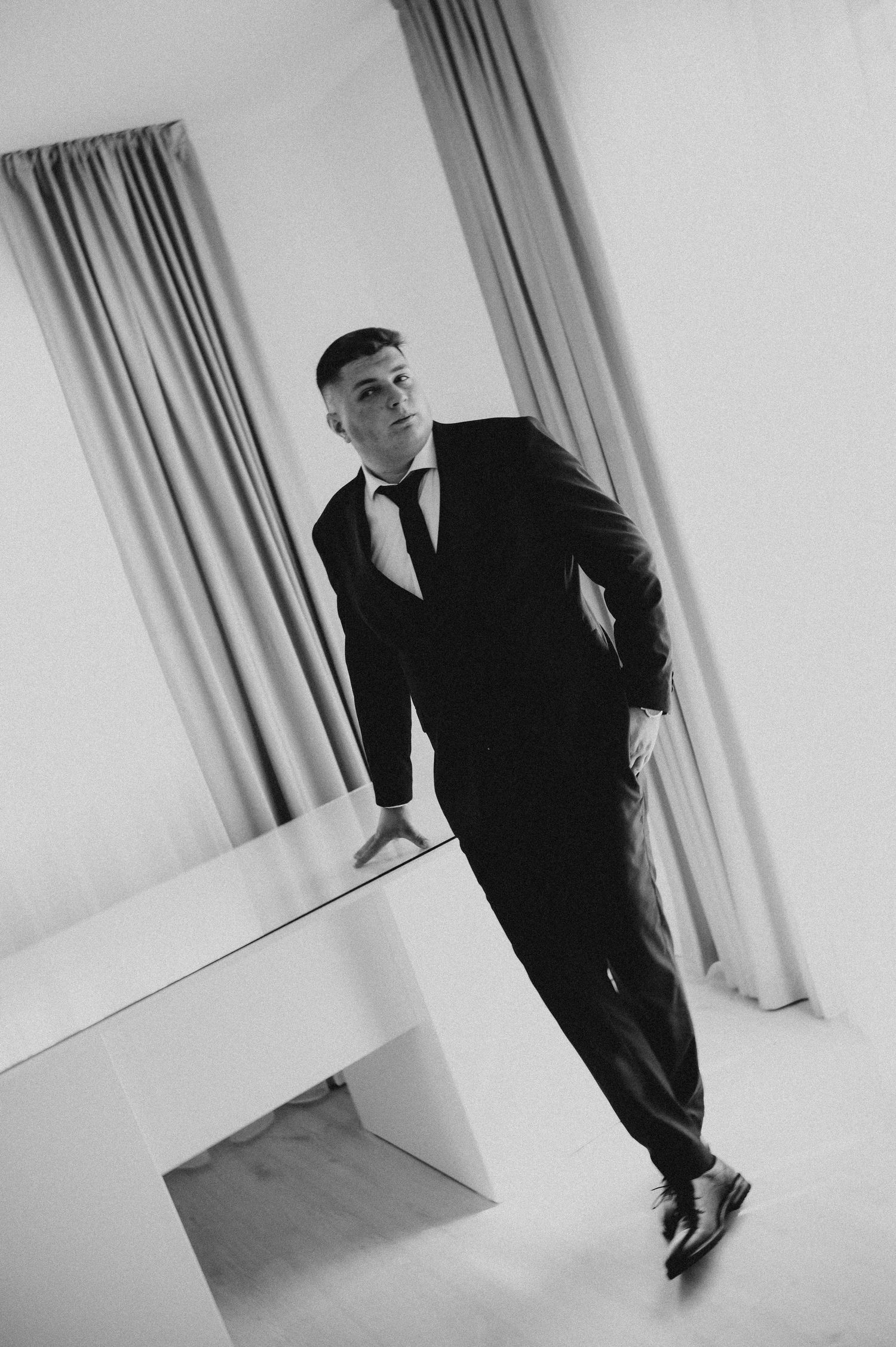 Elegant man in black tuxedo leaning against white table in minimalist room, black and white photo.