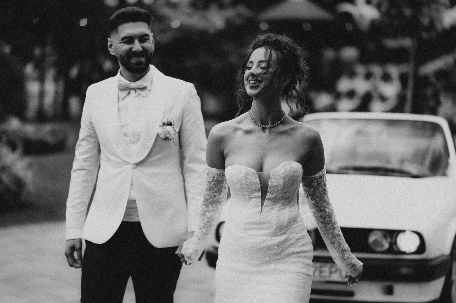 Black and white photo of a bride and groom holding hands, smiling, with a vintage white car in the background.