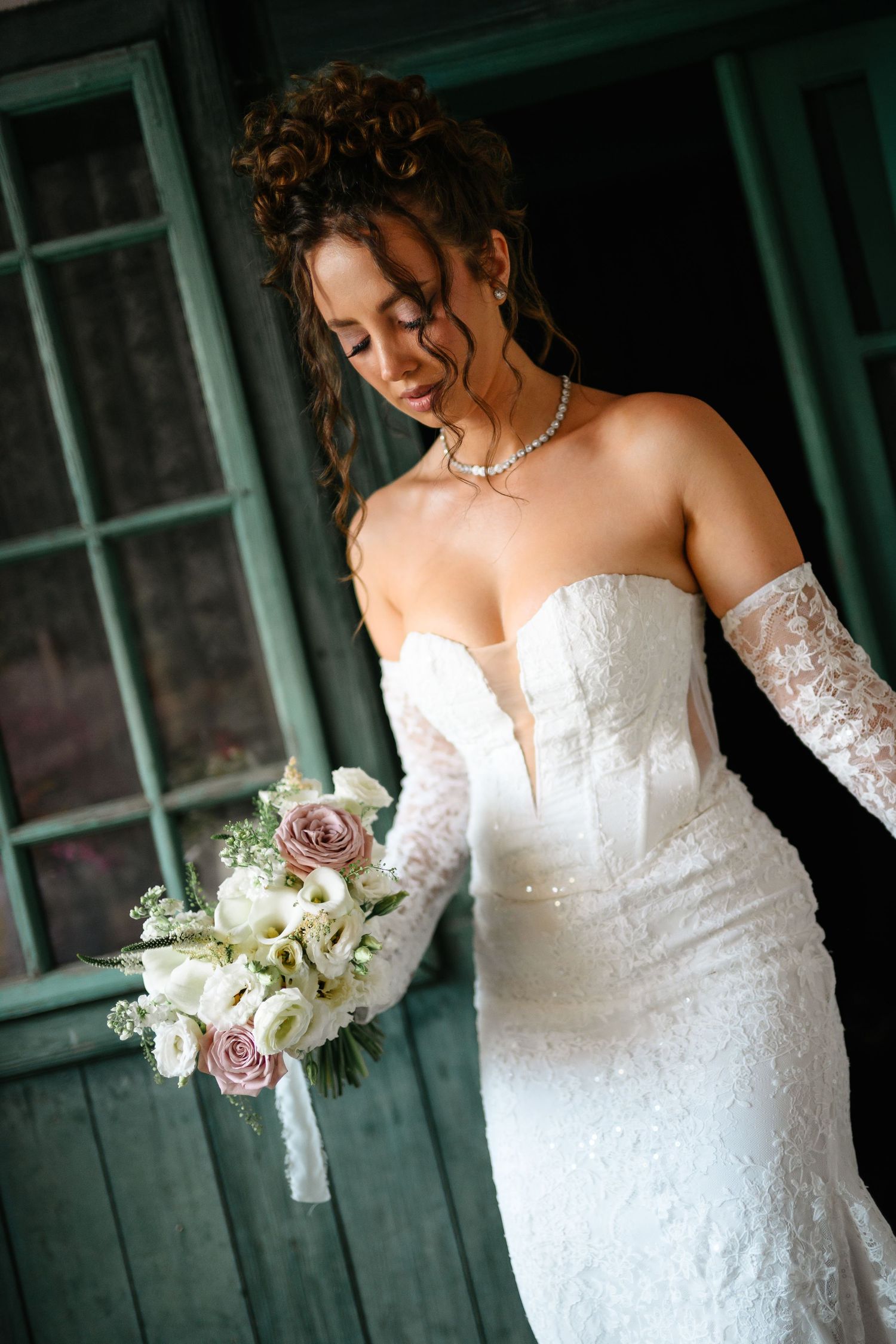 Elegant bride in white lace strapless gown holding a bouquet of pink and white roses, wearing pearl necklace.