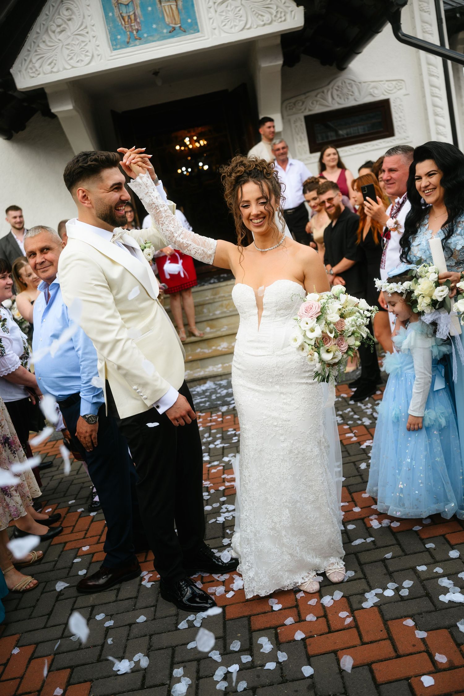 Smiling bride and groom celebrate outside church with confetti, guests applauding, bride holding white and pink bouquet.