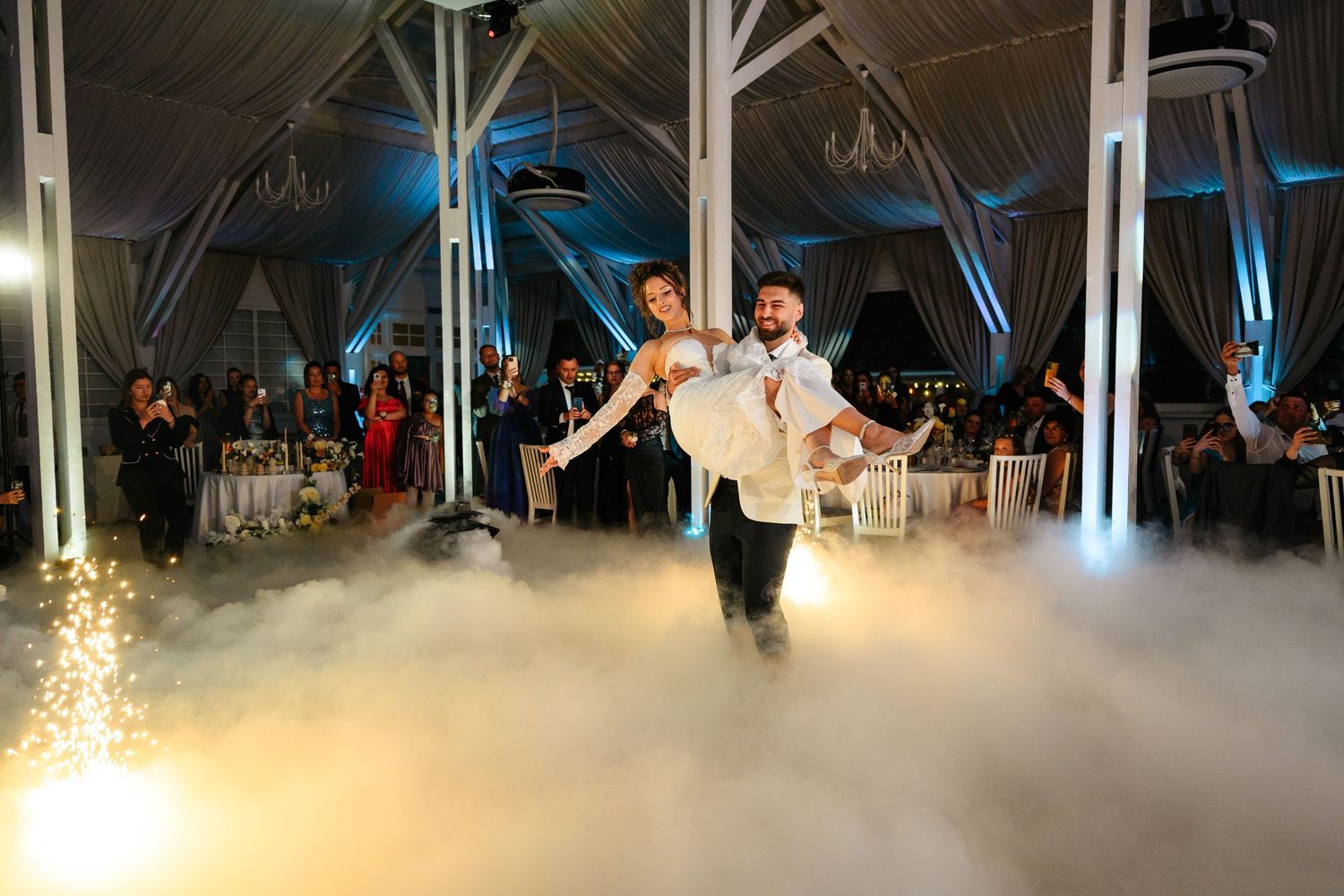 Groom lifting bride during first dance on foggy dance floor at elegant wedding reception.