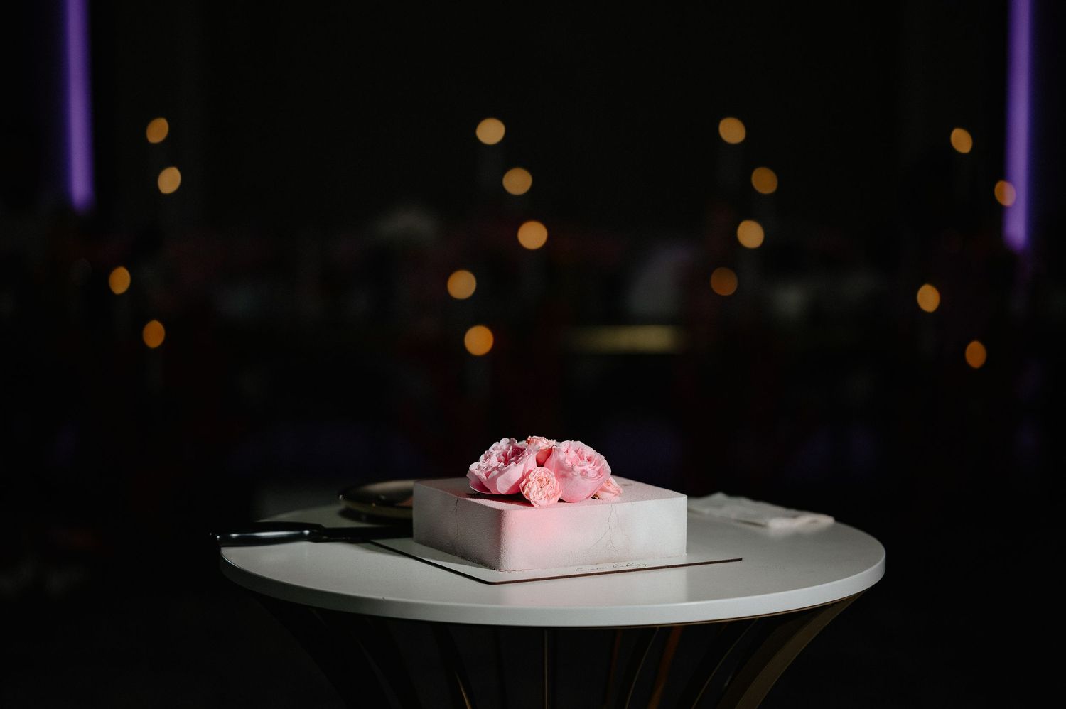 Elegant square white wedding cake topped with pink peonies displayed on a round pedestal table.