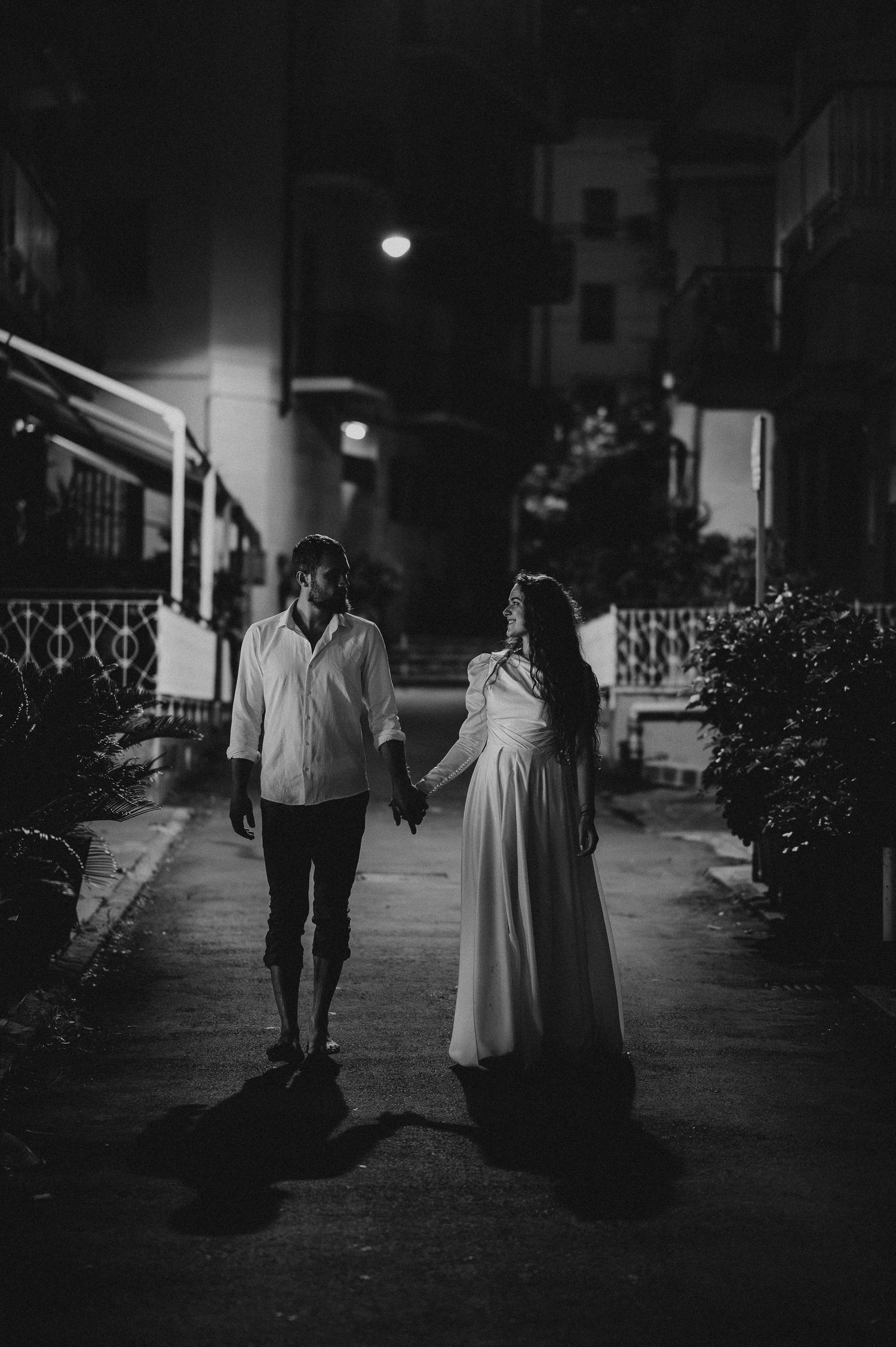 Black and white photo of a couple holding hands and walking along a quiet urban street at night.