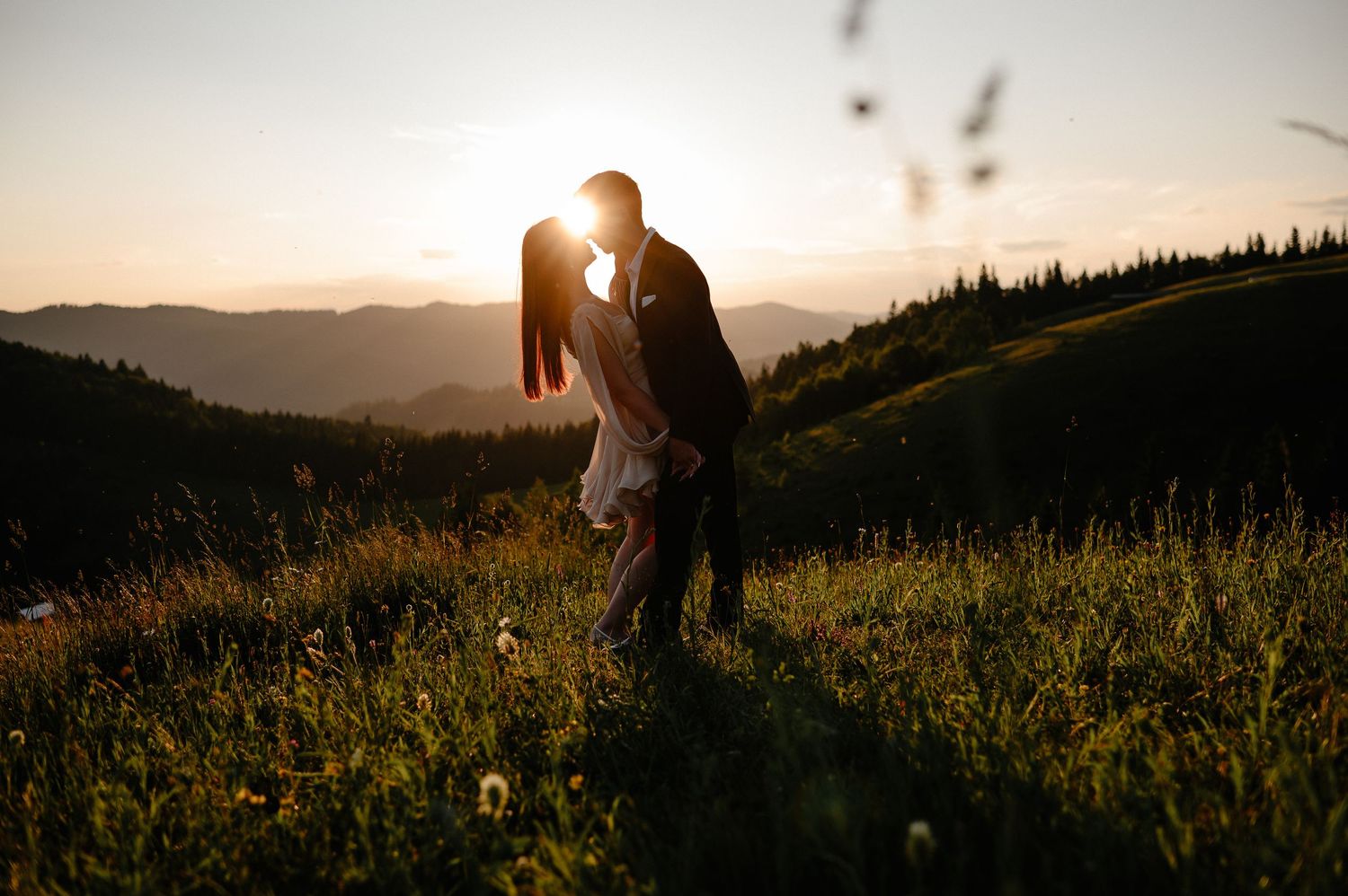 Romantic silhouette of couple kissing on a hillside meadow with stunning mountain sunset behind them.