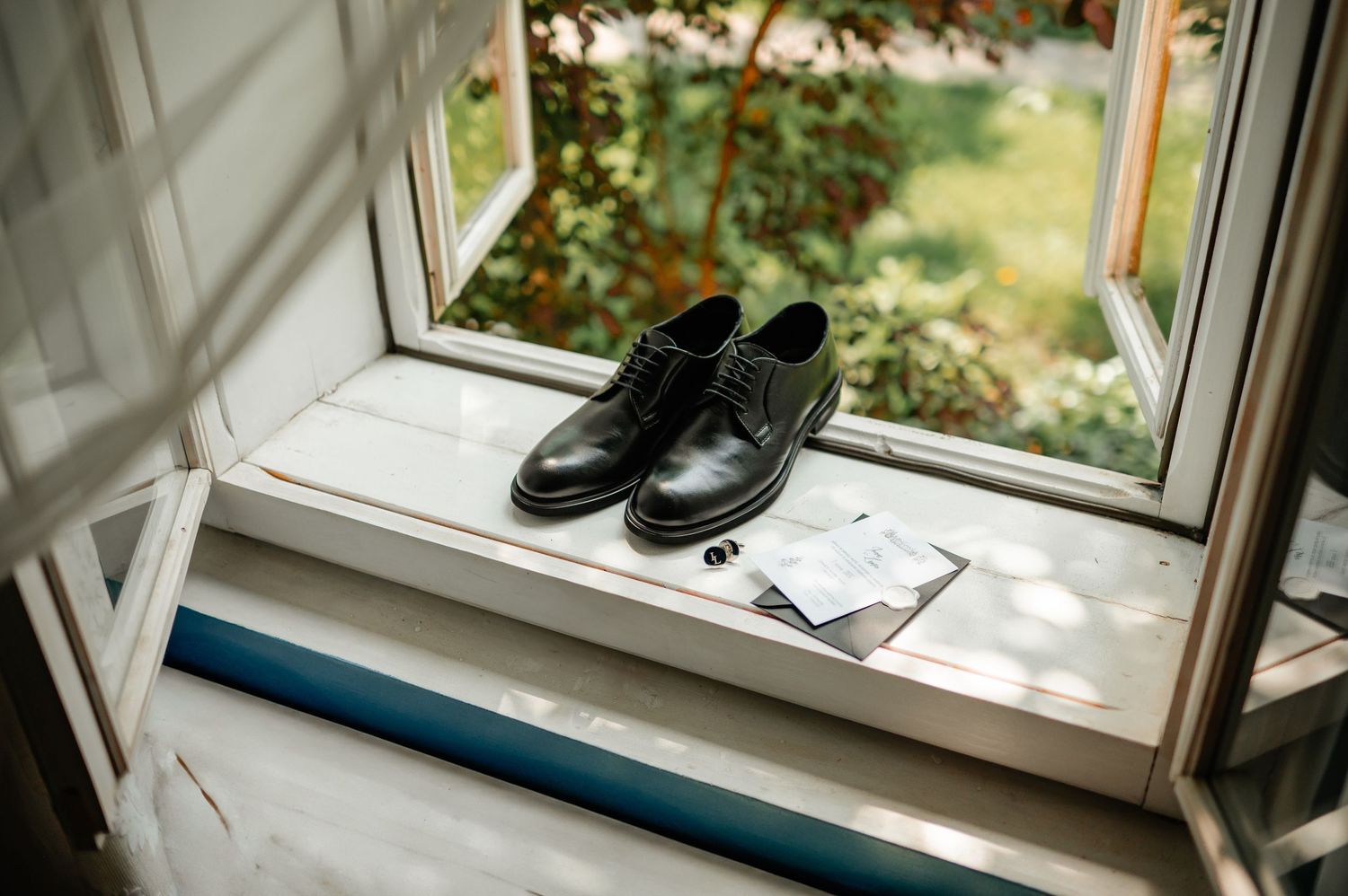 Classic black leather groom shoes with wedding invitation and cufflinks on white window sill.