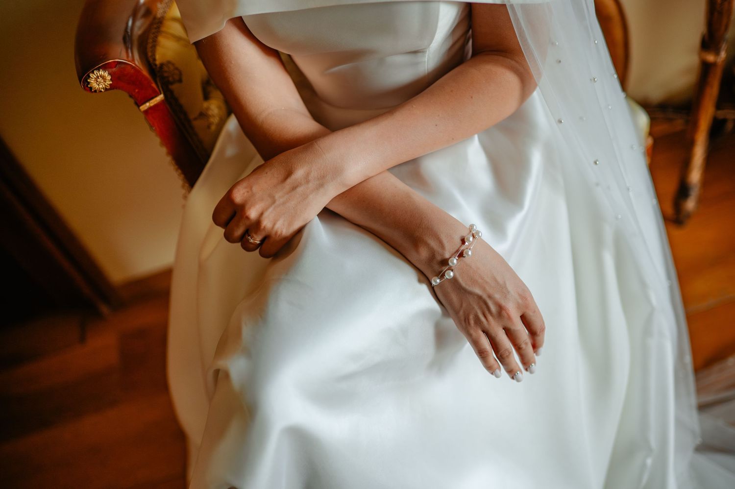 Close-up of bride in white satin wedding dress with delicate bracelet, hands folded elegantly.