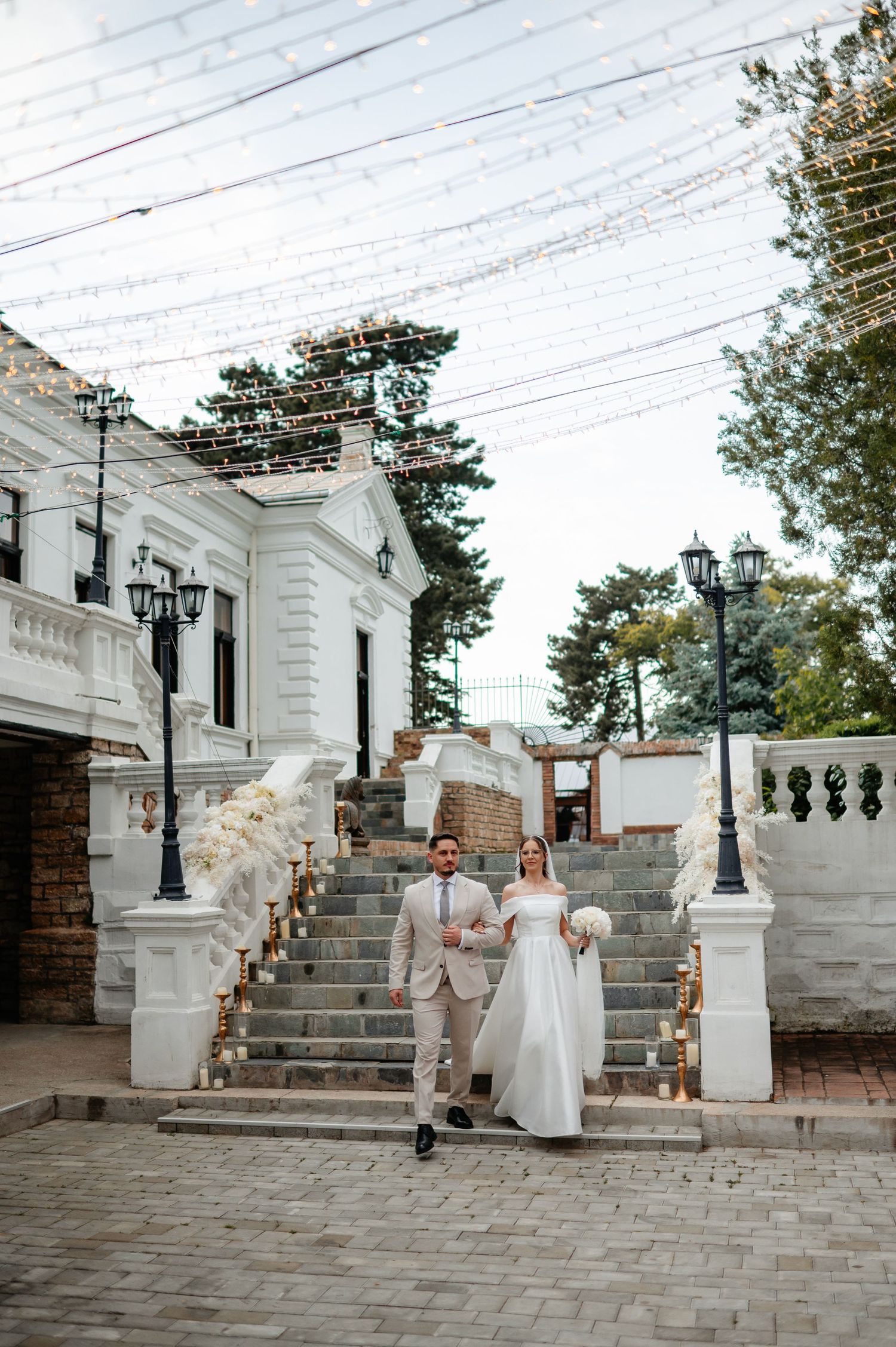Elegant bride and groom descend grand white staircase of neoclassical wedding venue under canopy of string lights.