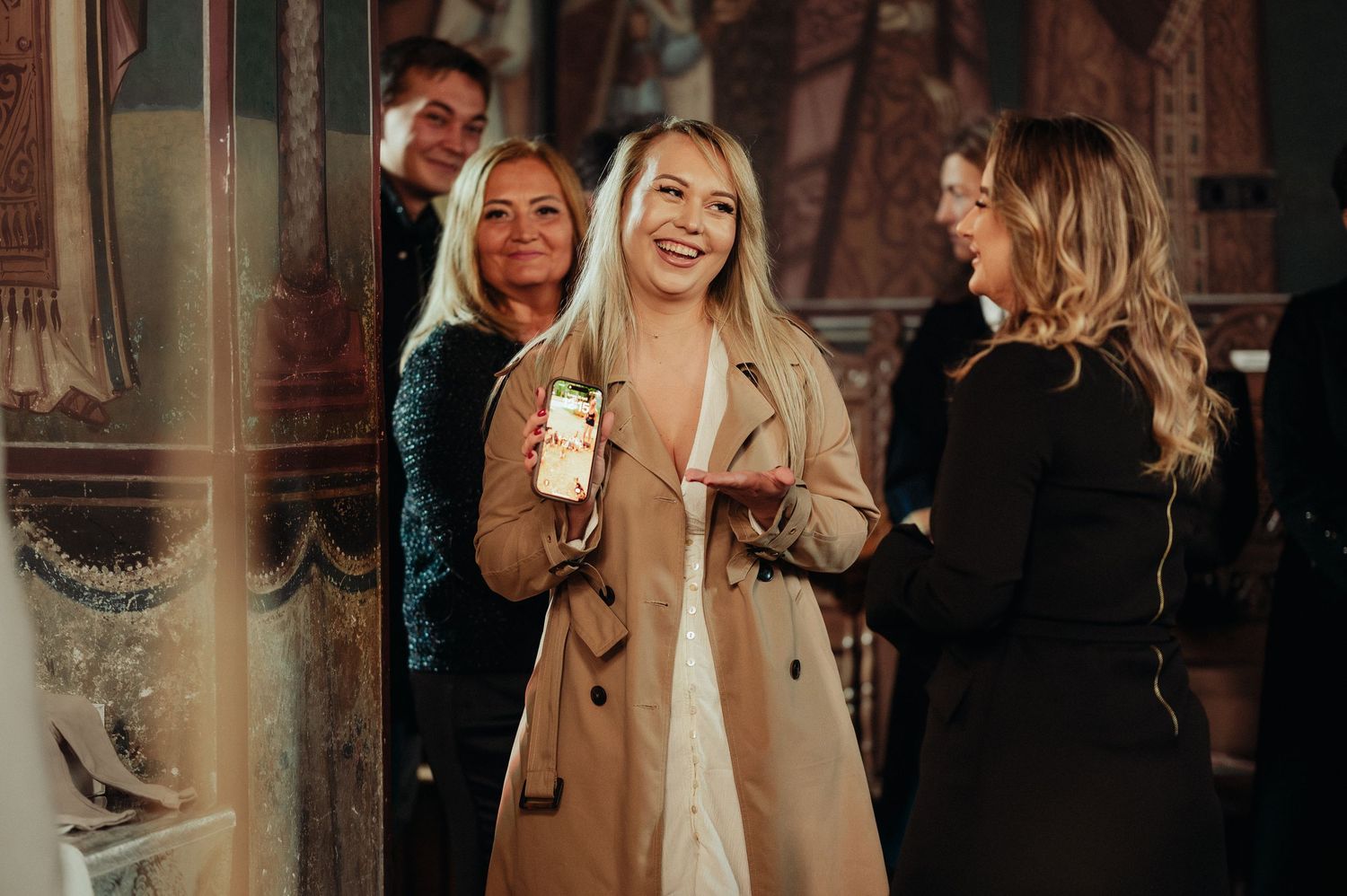 Smiling blonde woman in beige coat holding phone at elegant indoor social gathering.