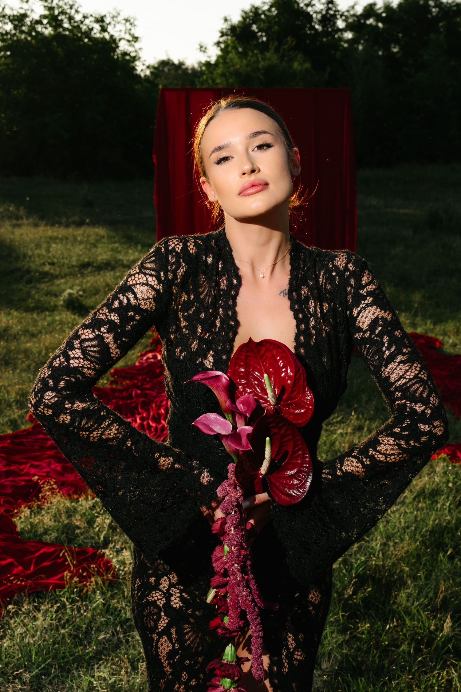 Woman in black lace dress holding red anthurium flowers, posing outdoors on grass with red fabric backdrop.