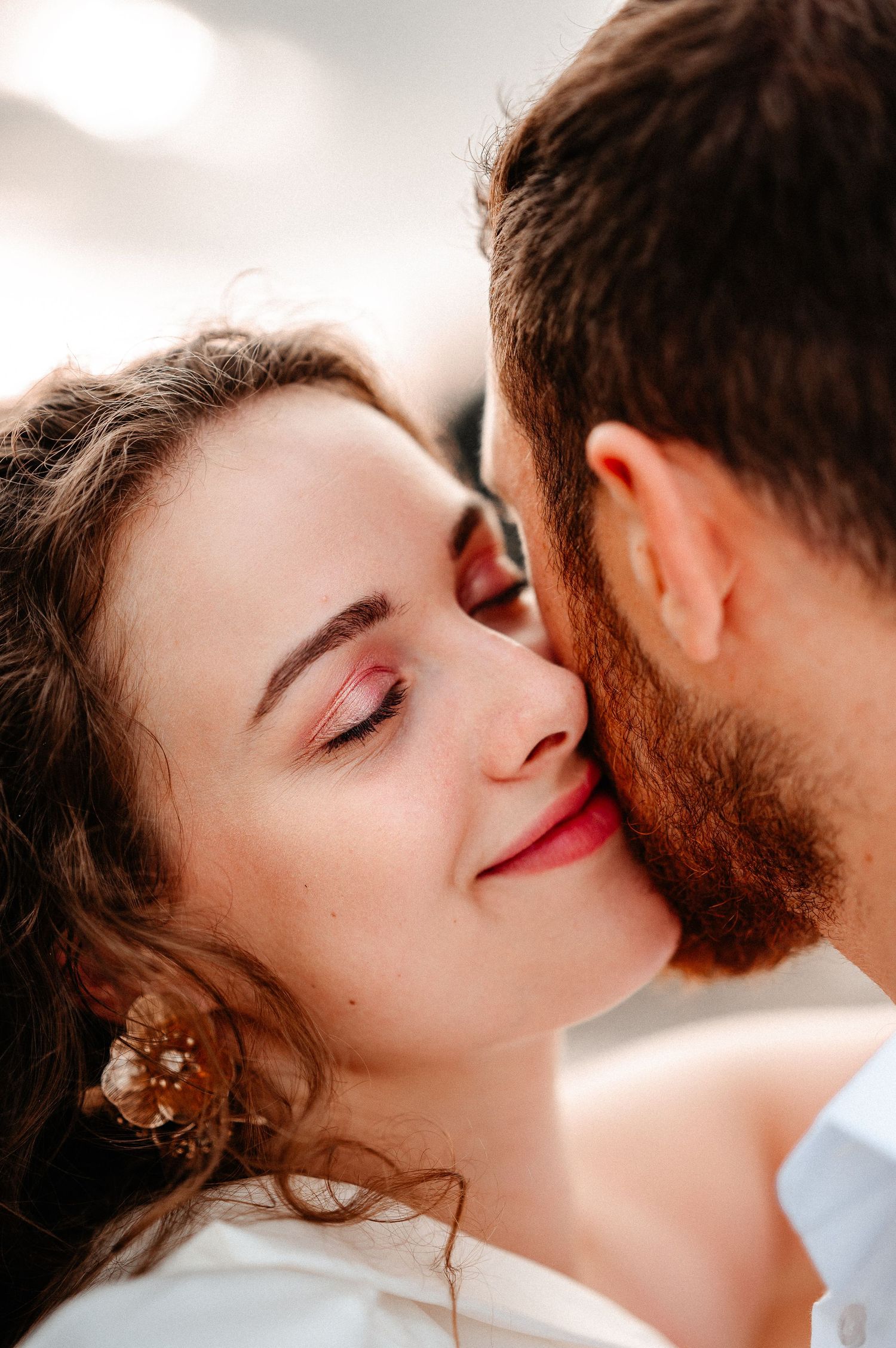 A bearded man gently kisses the cheek of a smiling young woman with curly hair and pink earrings.