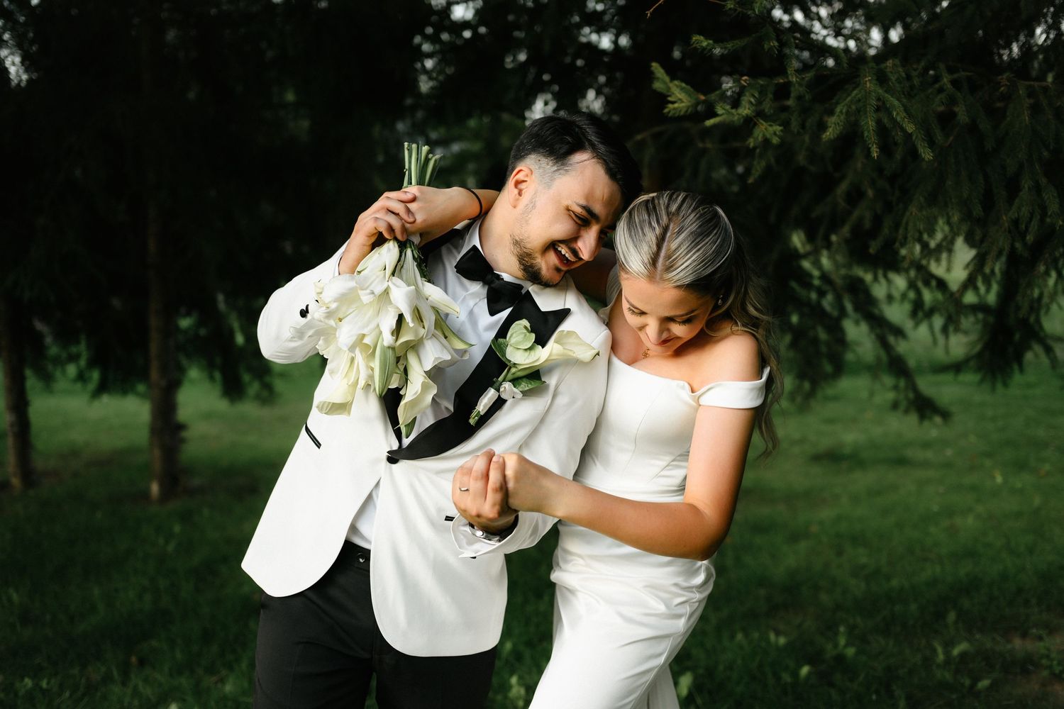 A joyful bride and groom share a playful moment outdoors, with the groom holding a white floral bouquet.