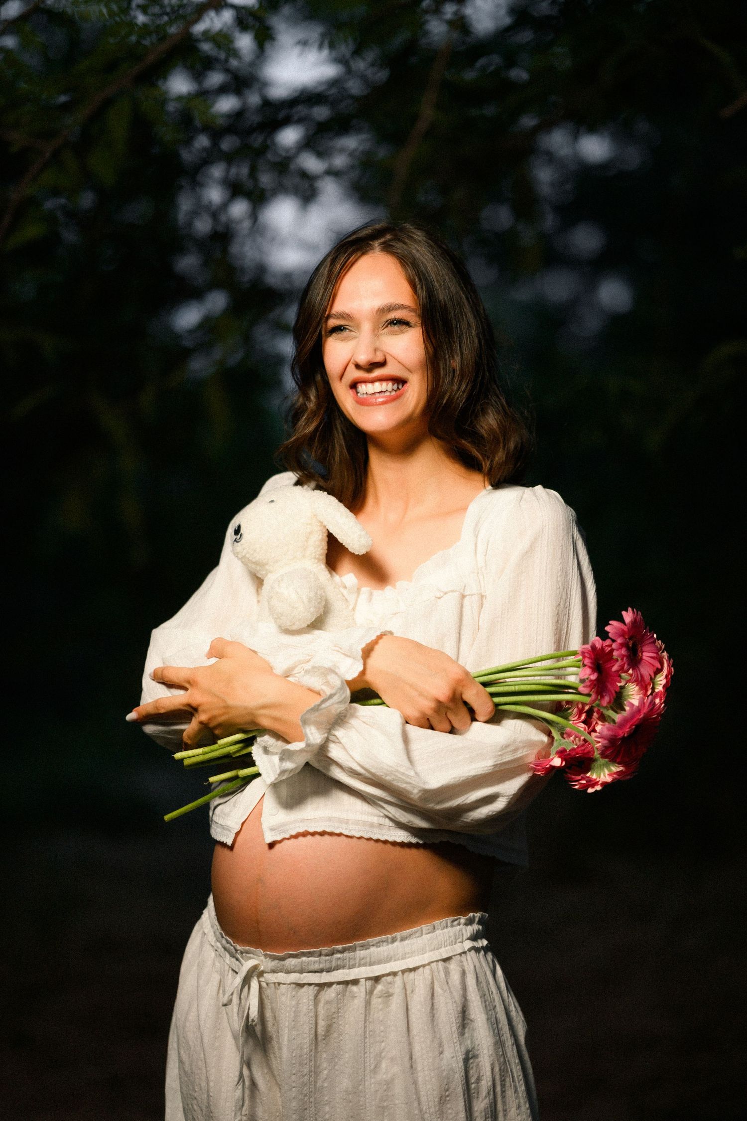Smiling pregnant woman holding a white teddy bear and pink gerbera flowers outdoors in natural light.