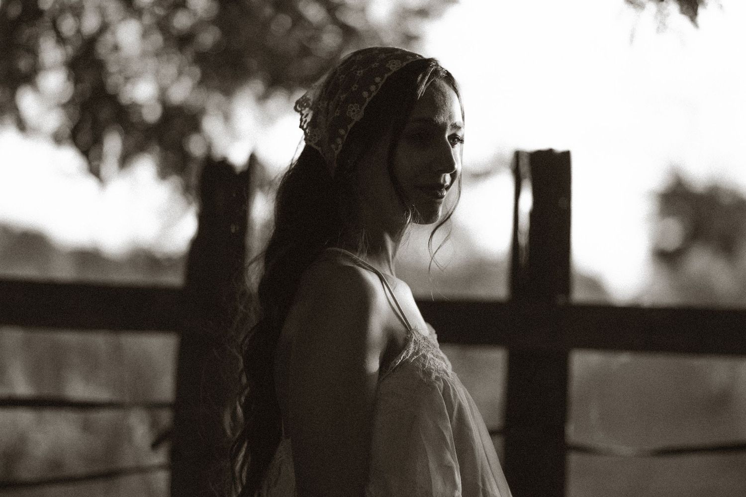 Black and white portrait of a young woman with braided hair in a flowy dress standing near a wooden fence.