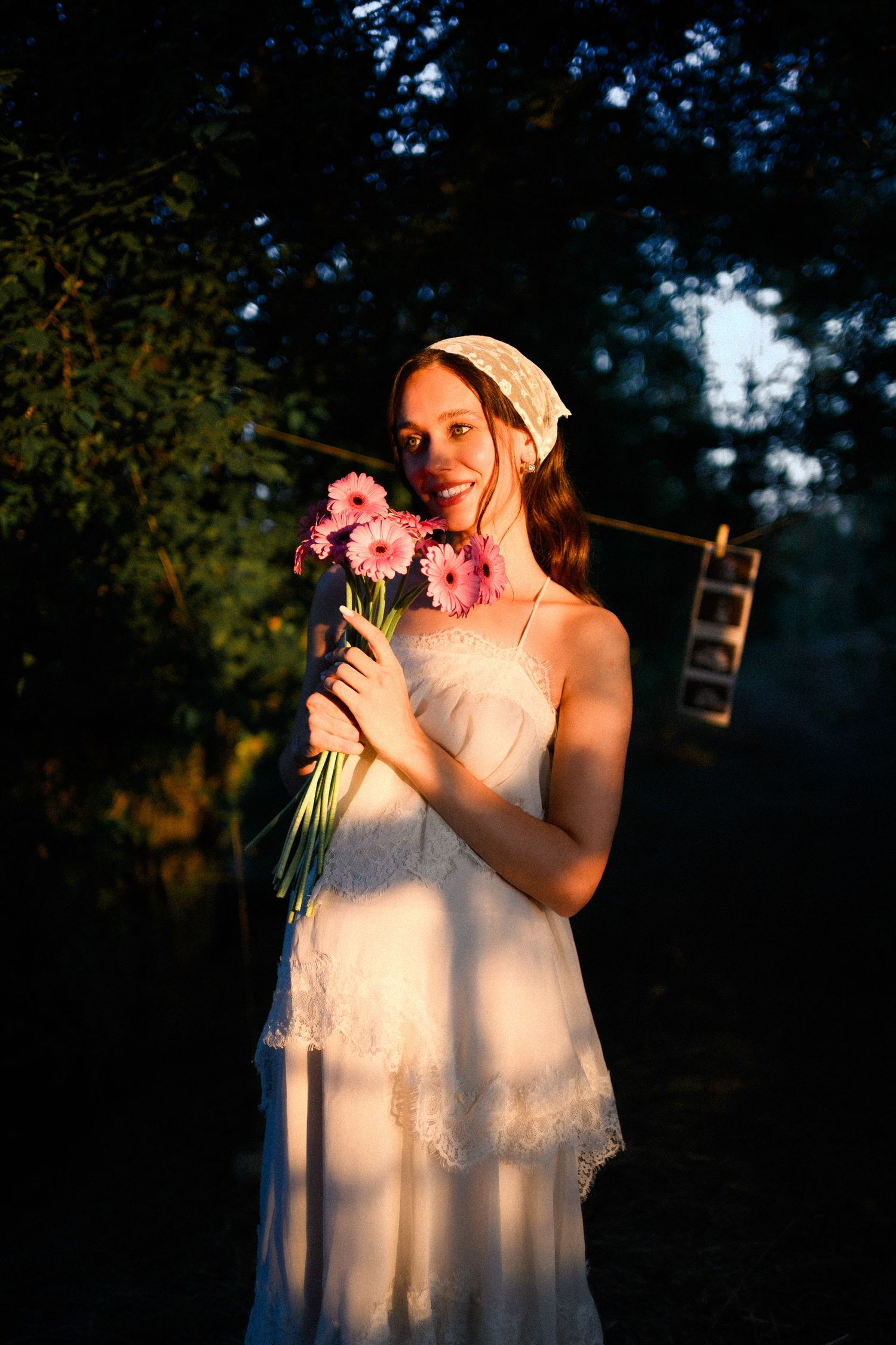 Young woman in white lace dress holding pink gerbera daisies, smiling in golden sunset light outdoors.