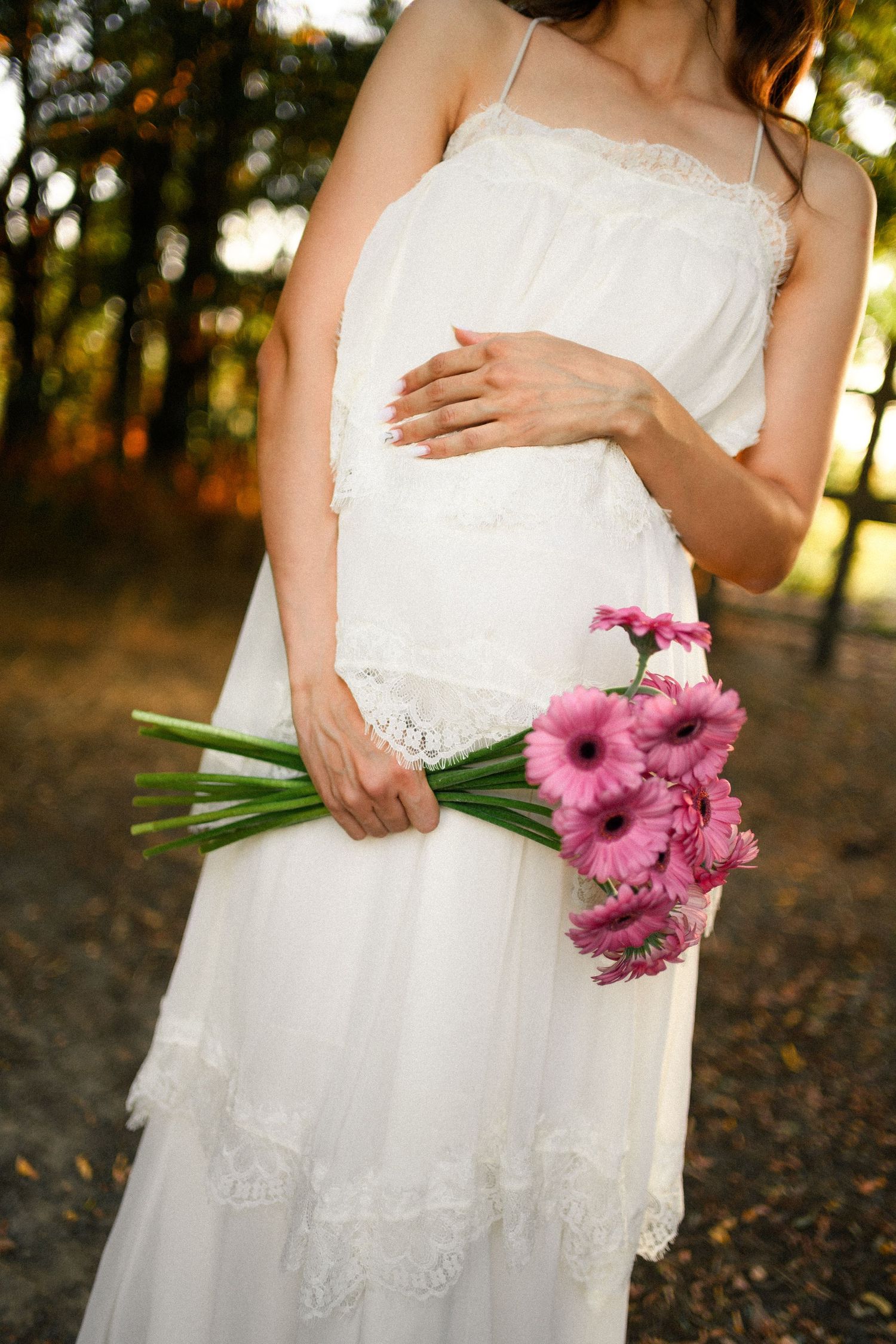 Pregnant woman in white lace dress holding pink gerbera daisy bouquet outdoors in a sunlit forest setting.