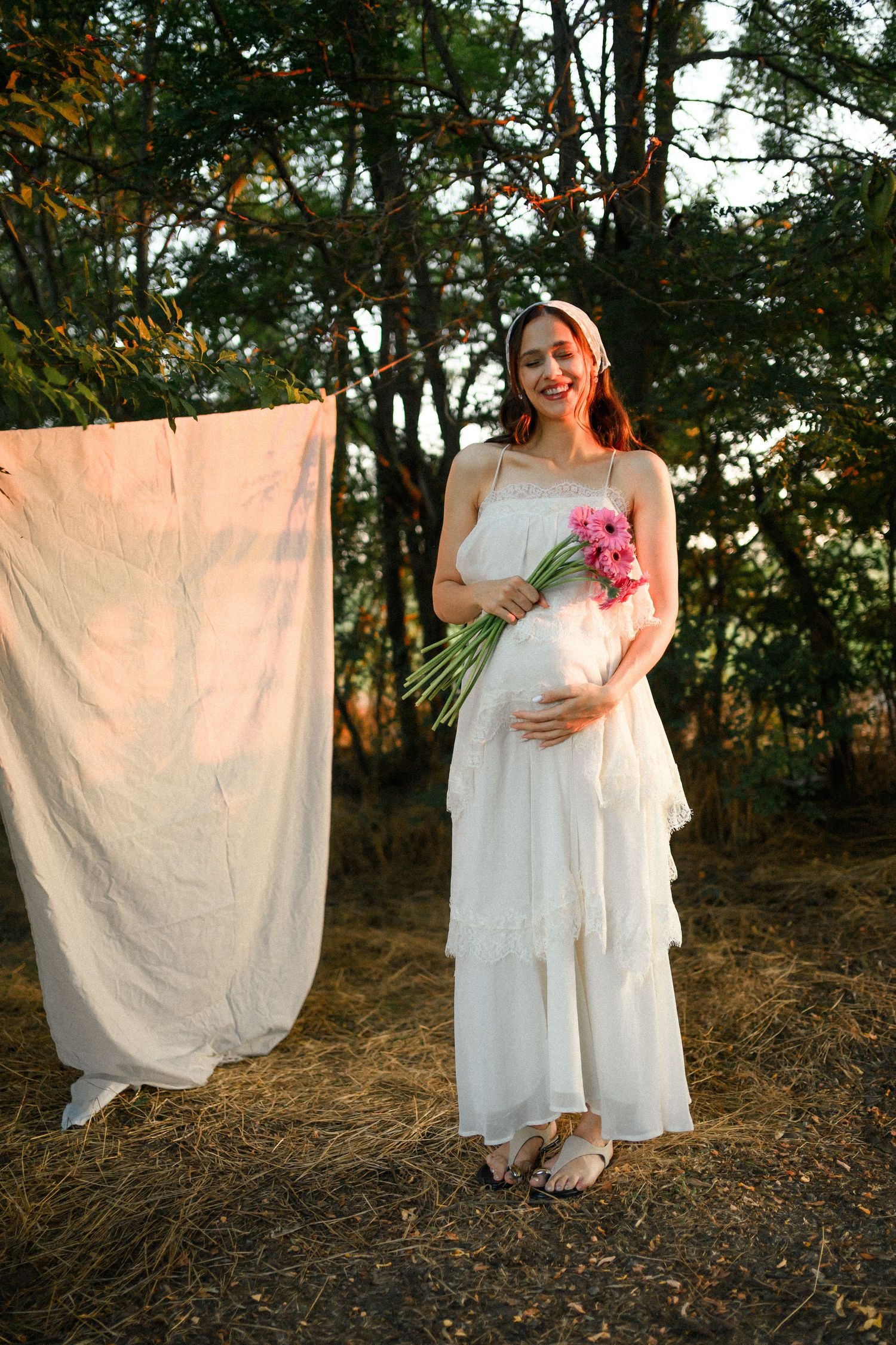 Smiling pregnant woman in white dress holding pink flowers, standing outdoors near hanging linen and trees at sunset.
