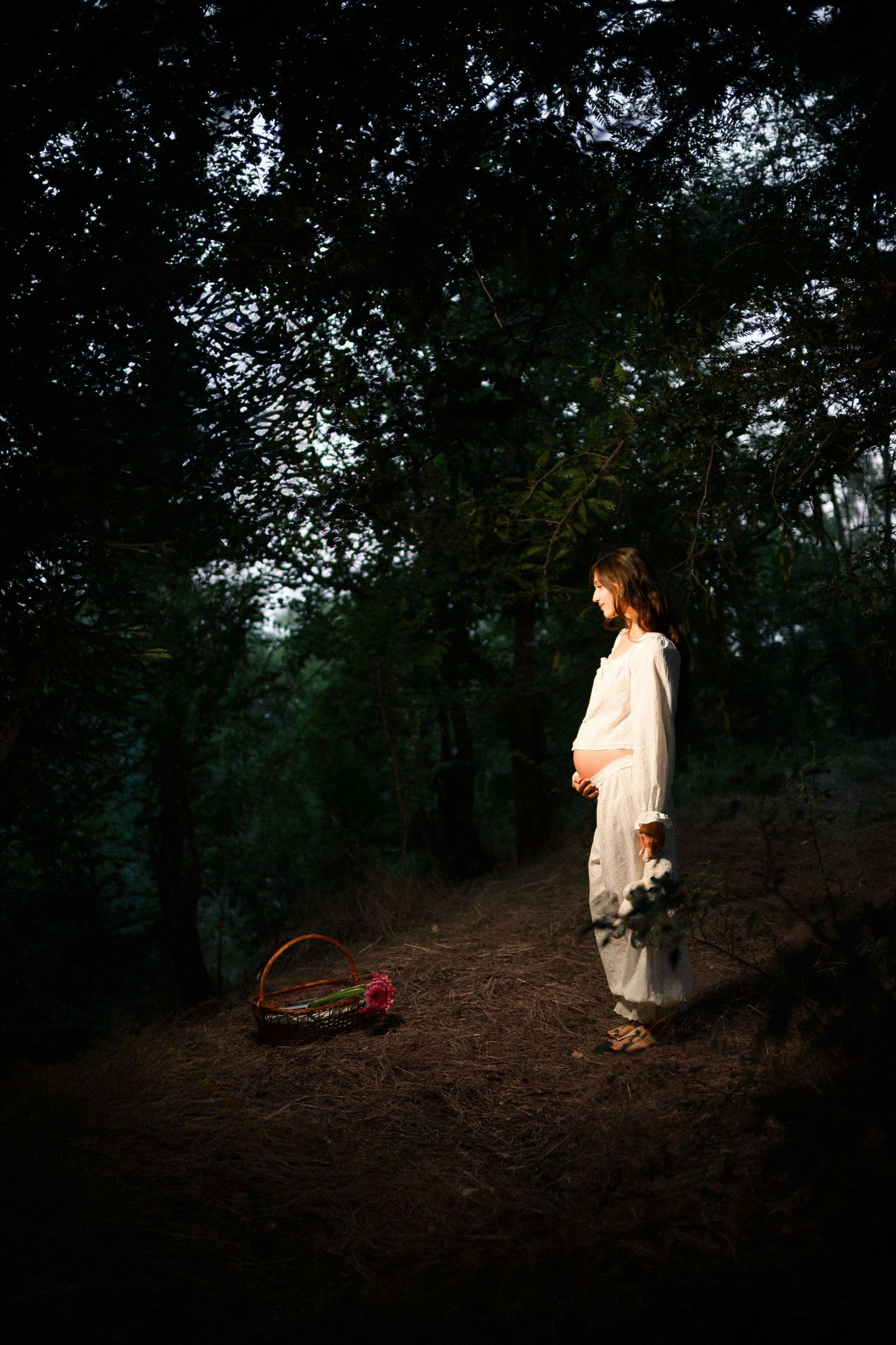 Young woman in white dress standing on forest path at dusk, holding flowers beside a wicker basket.