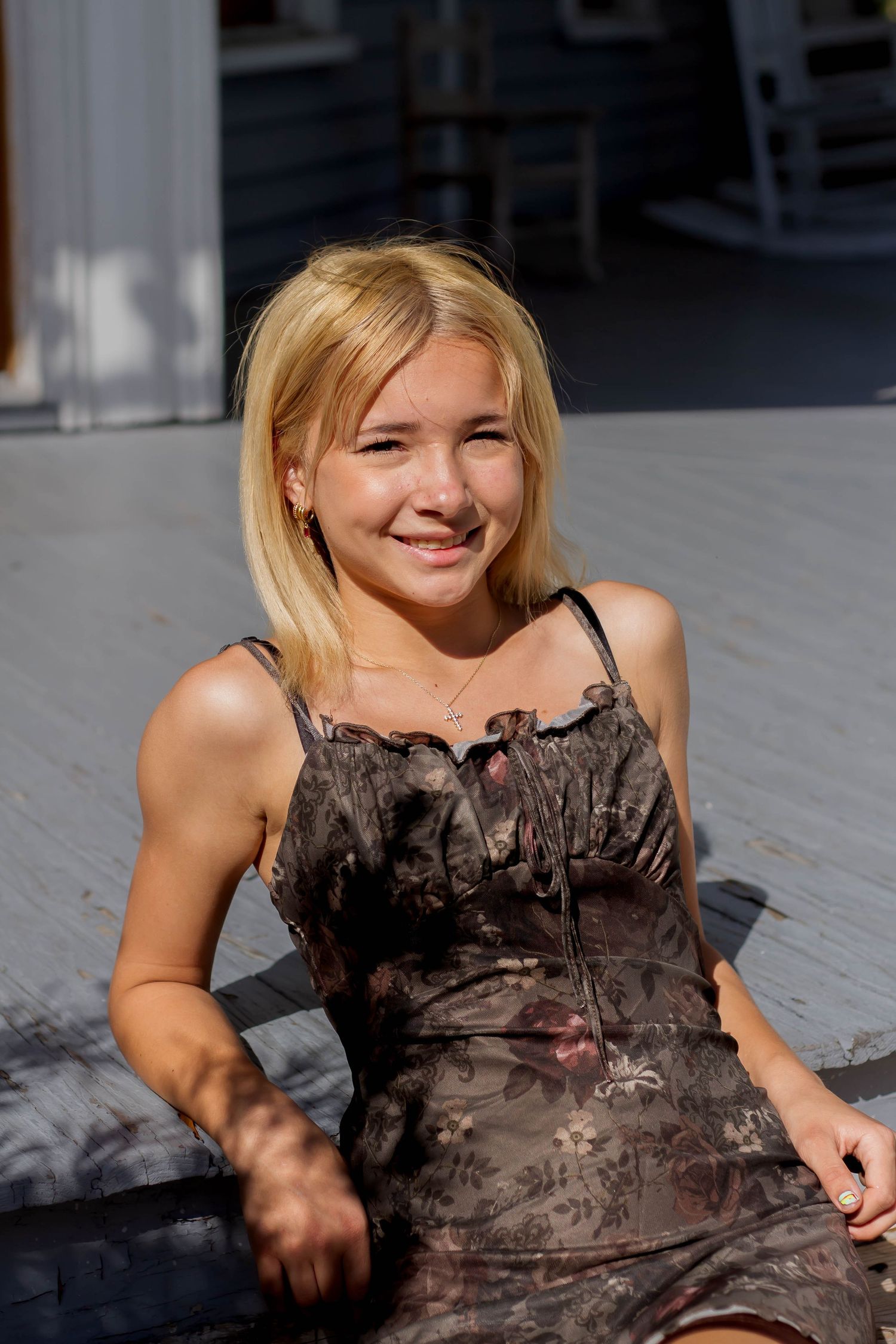 Smiling young blonde woman in dark floral dress sitting on sunny outdoor wooden steps.