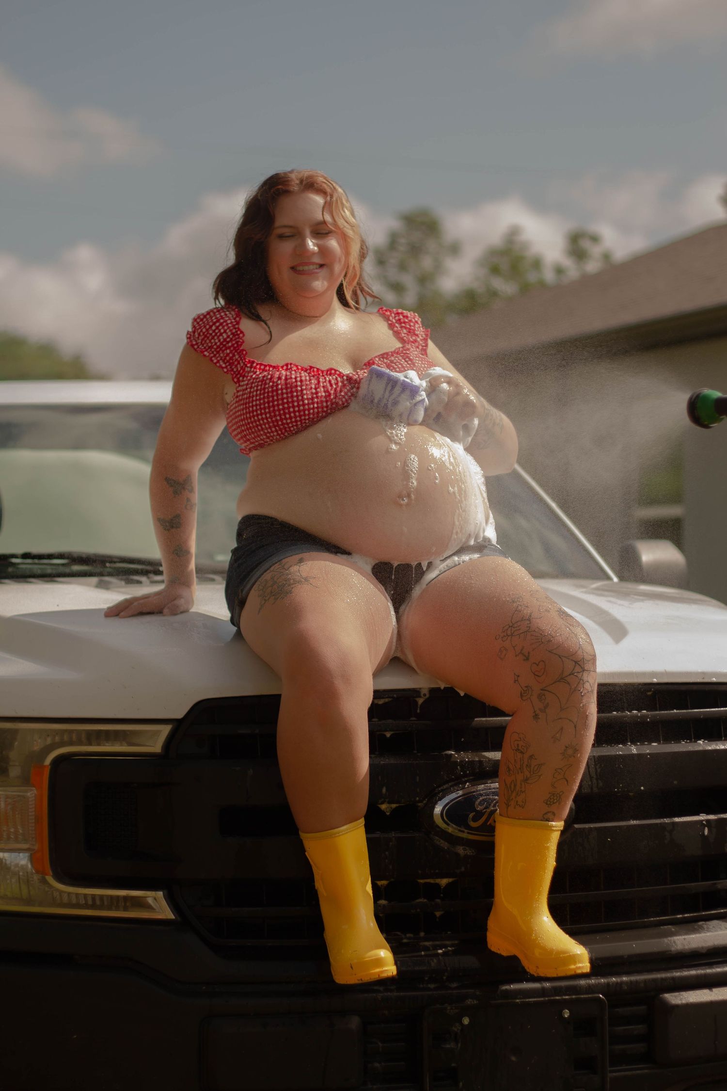 A pregnant woman in a red gingham top and yellow boots sits on a Ford truck hood while washing it.