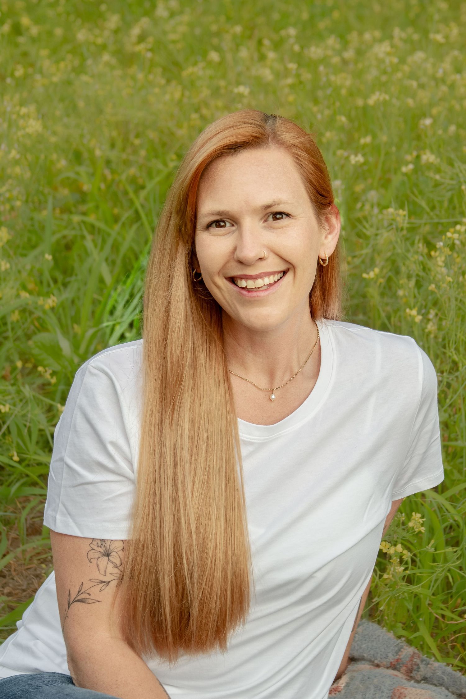 Smiling redhead woman with long straight hair and tattoo in white tee sitting outdoors.