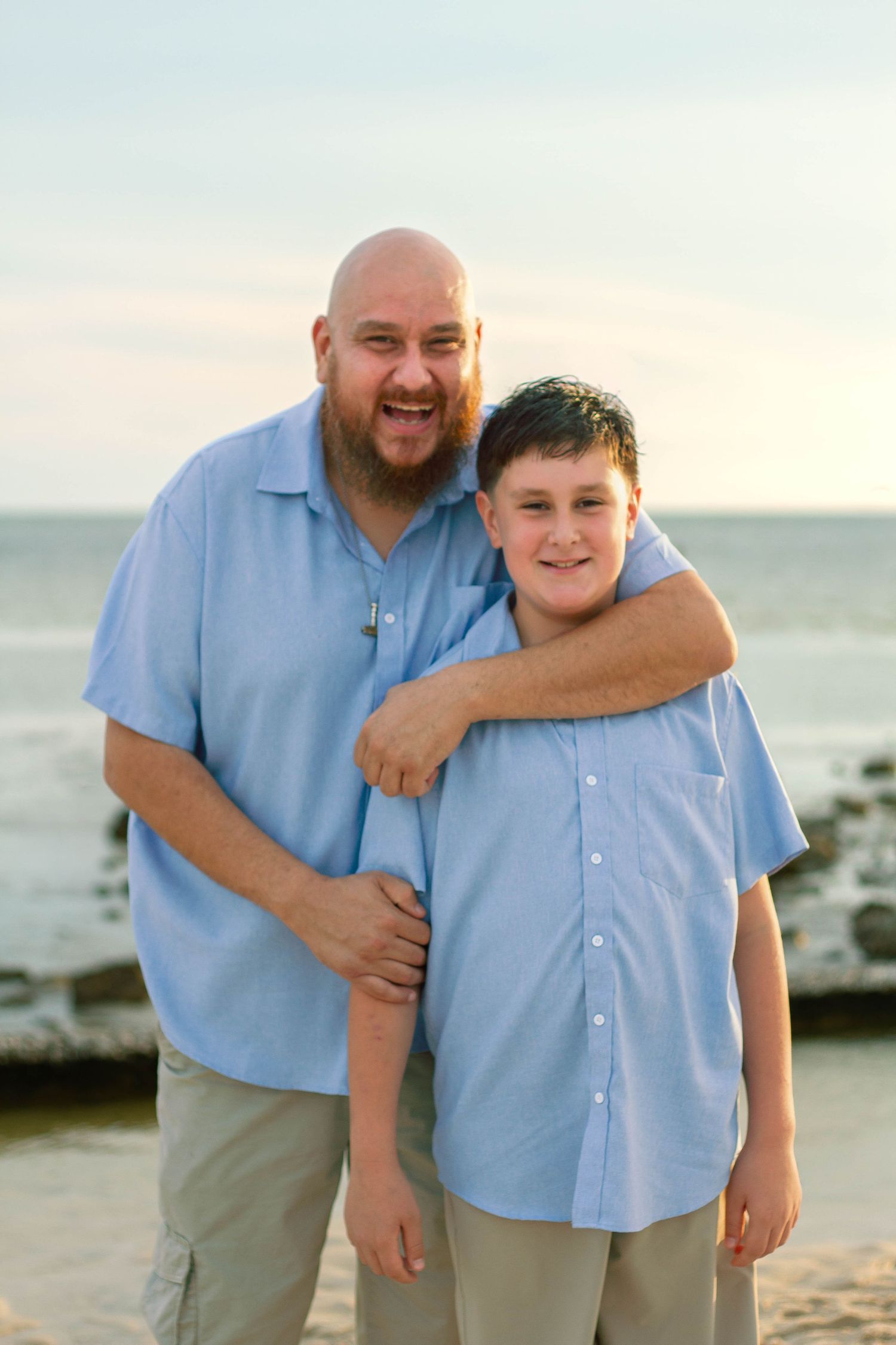 Laughing bearded man hugs smiling young son on a sandy beach during a family photo session.