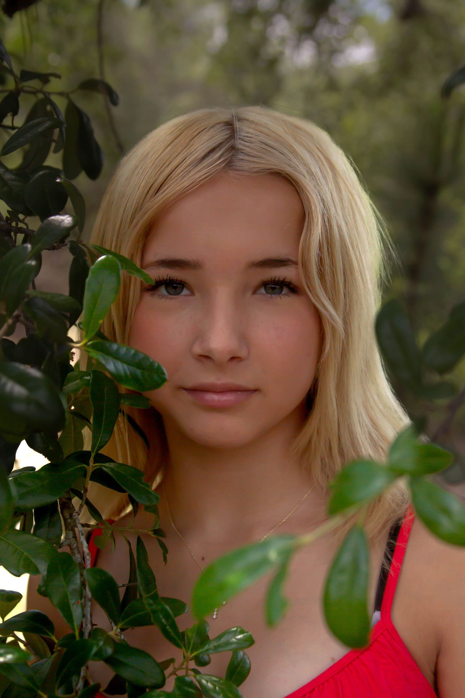 Young blonde woman partially framed by green leaves wearing a red dress in a natural setting.
