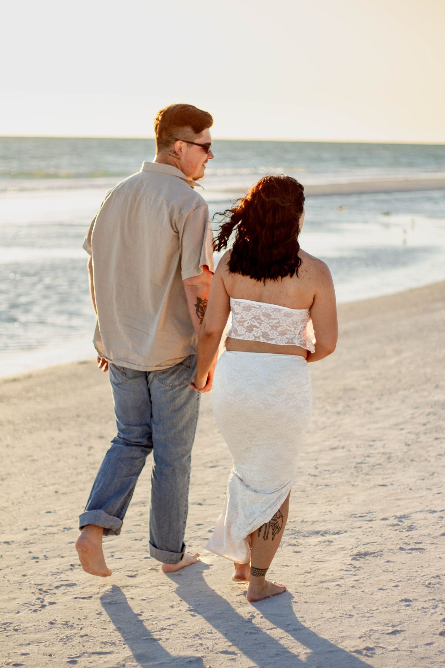 Couple walking hand in hand on sandy beach at sunset from behind.