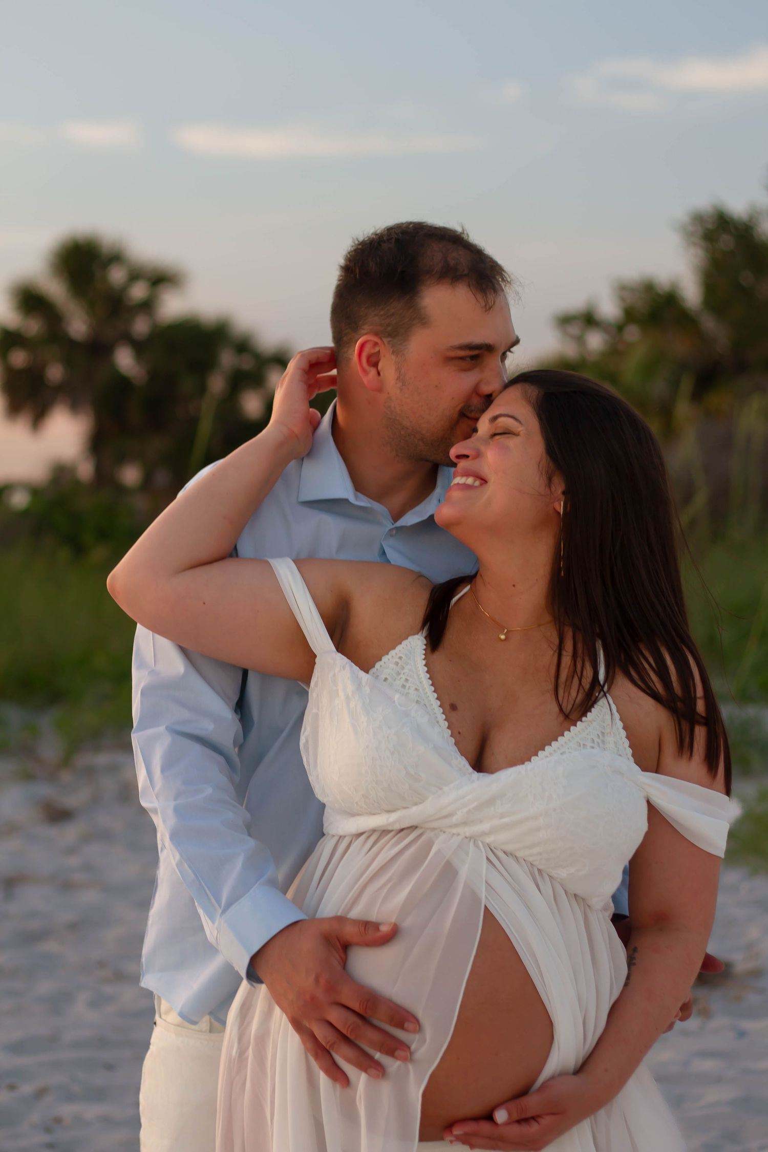 Man kisses pregnant woman's cheek from behind as they stand together on a sandy beach at dusk.