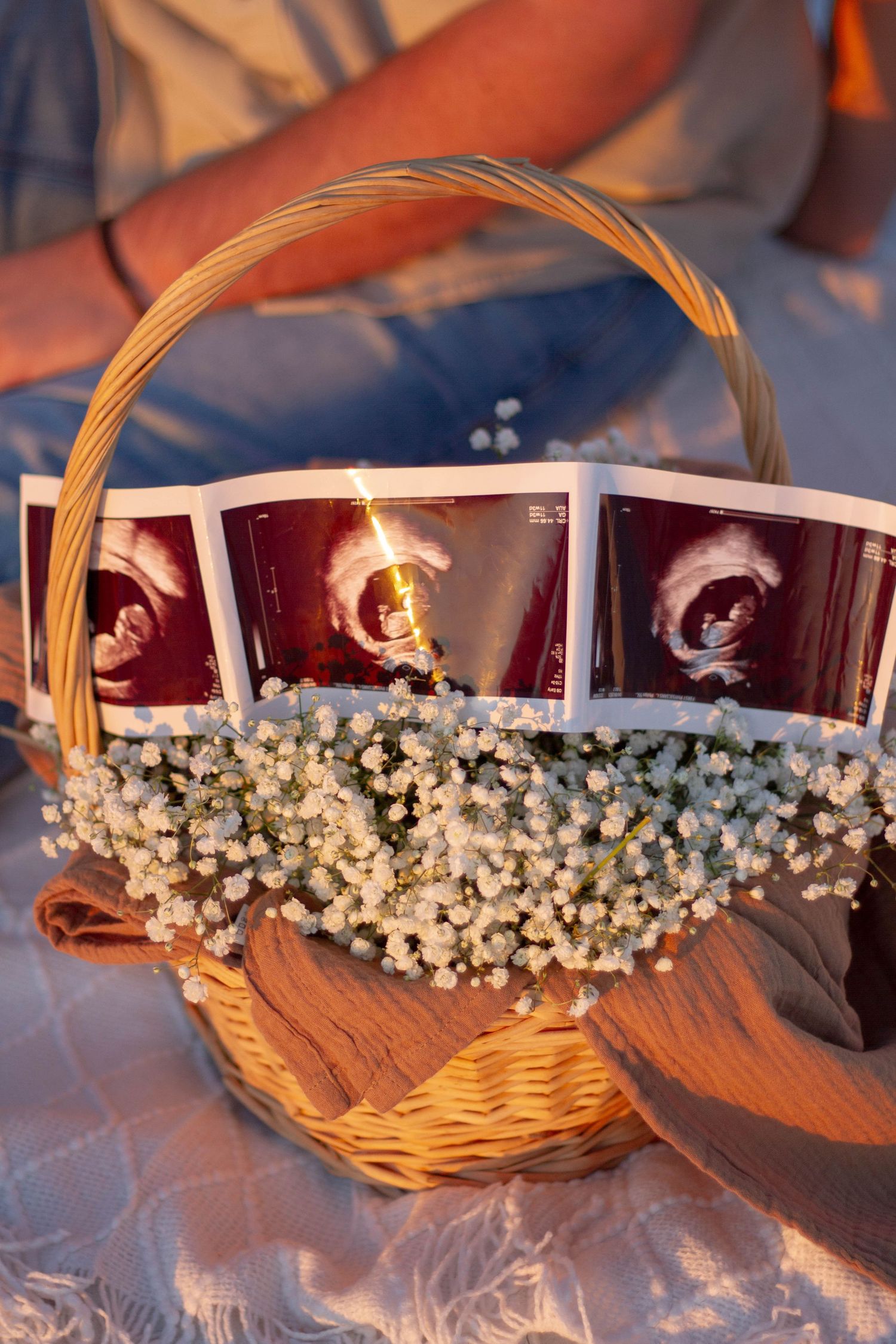 Wicker basket decorated with baby's breath flowers displaying ultrasound photos on a soft blanket outdoors.