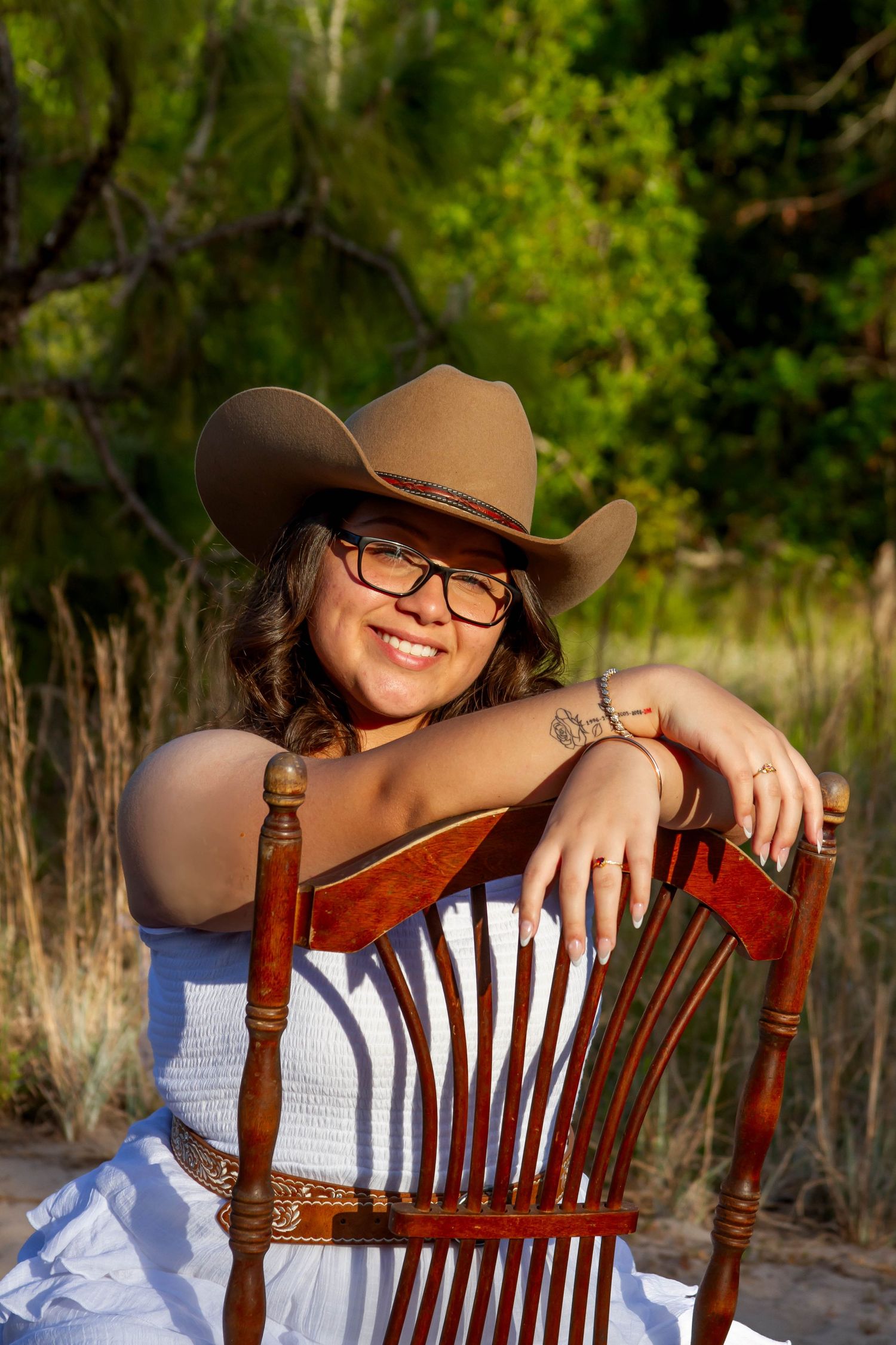 Smiling young woman in cowboy hat sitting on wooden chair in sunny outdoor setting.