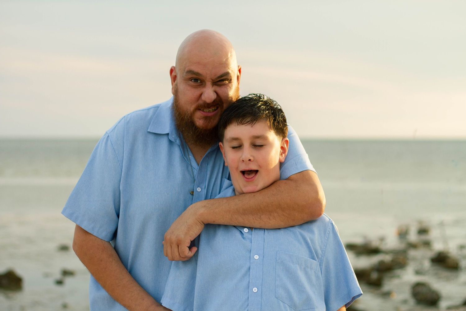 Bearded father playfully holds teenage son in headlock during family beach photo session.
