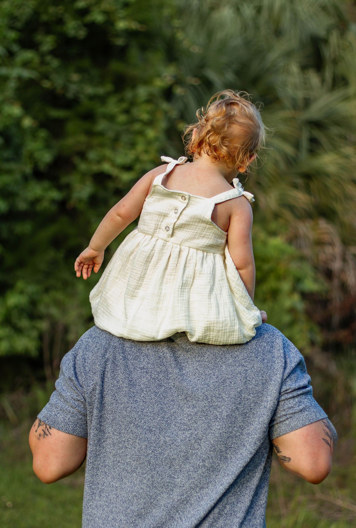 A toddler in a yellow dress sits on an adult's shoulders outdoors with green trees in the background.