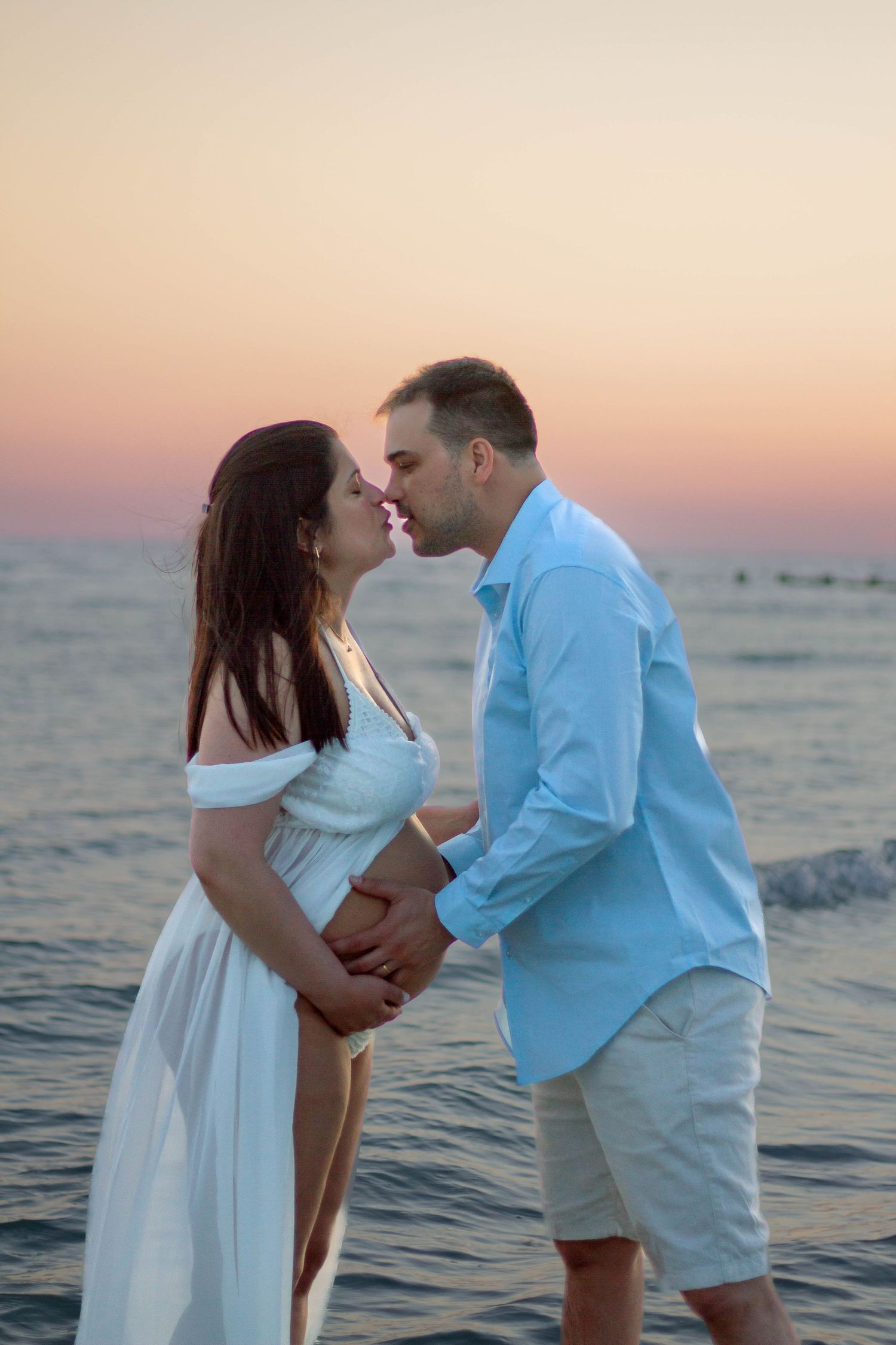 Pregnant couple kissing at sunset on beach near ocean.