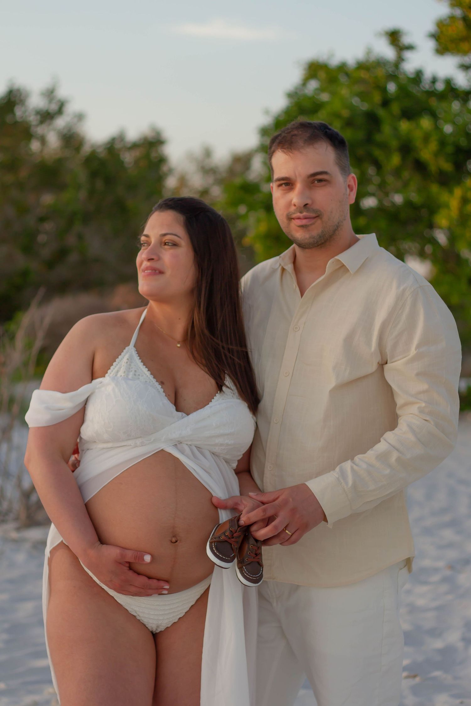 Pregnant couple posing on beach, man holding tiny baby shoes while embracing woman in white outfit and flowing wrap.