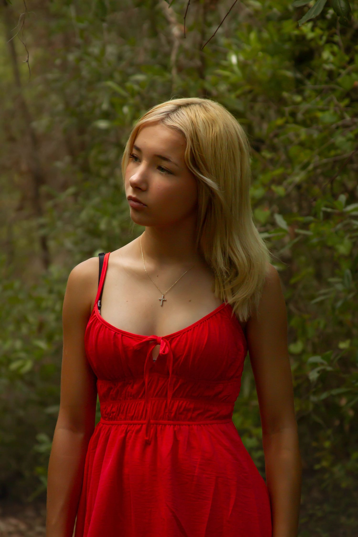 Young blonde woman in red sundress with cross necklace standing outdoors among green trees.