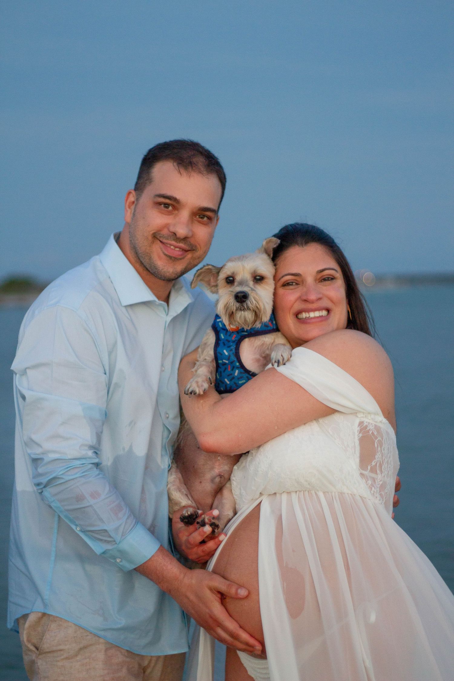 Smiling pregnant couple holding small dog in blue bandana near waterfront during maternity photo session.