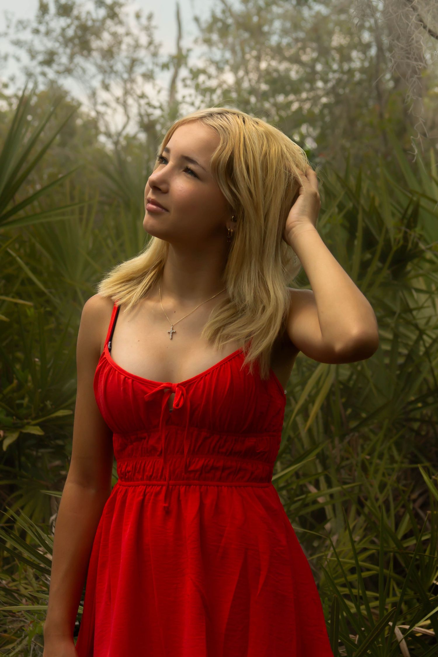 Blonde woman in red sundress with cross necklace posing among palm plants in a misty forest.