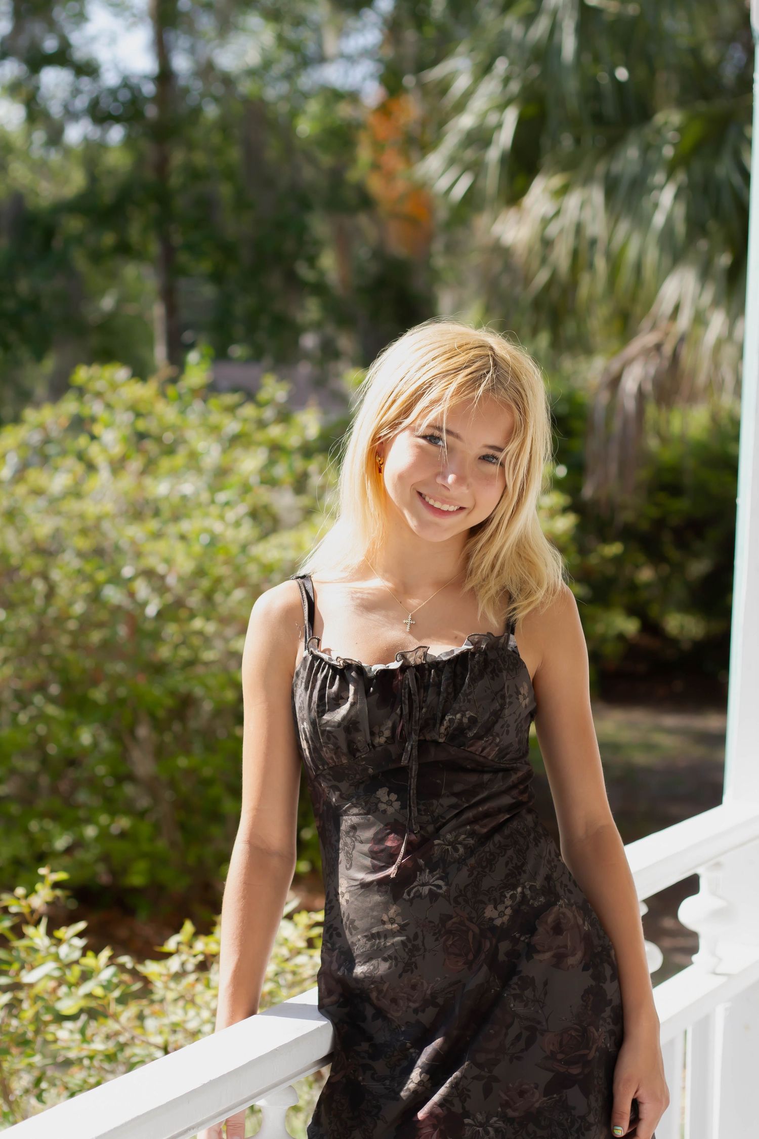 Smiling blonde woman in a dark floral dress standing on a sunny porch with green trees behind.
