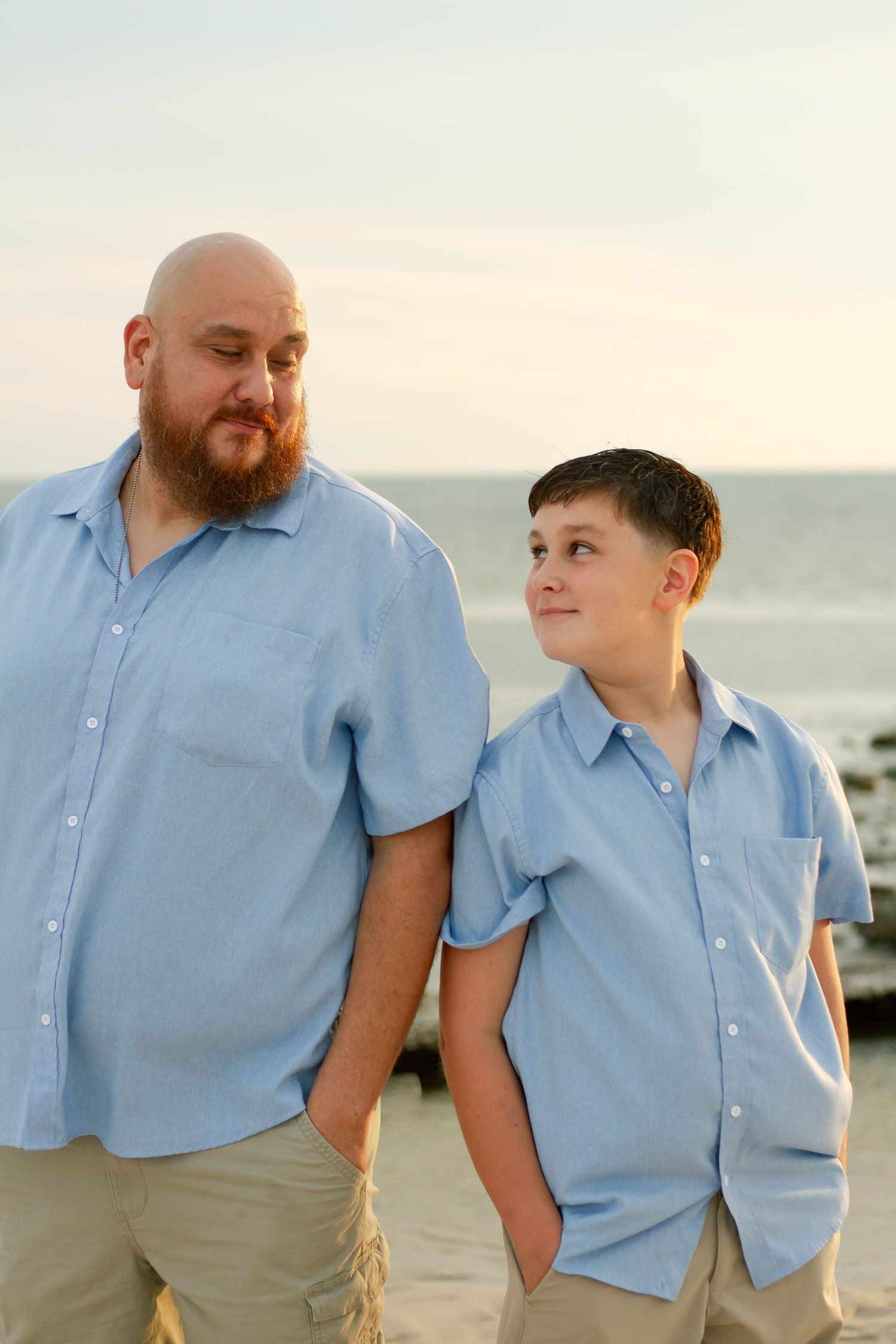 Bearded man in blue shirt stands with arm around young son near the ocean at sunset.