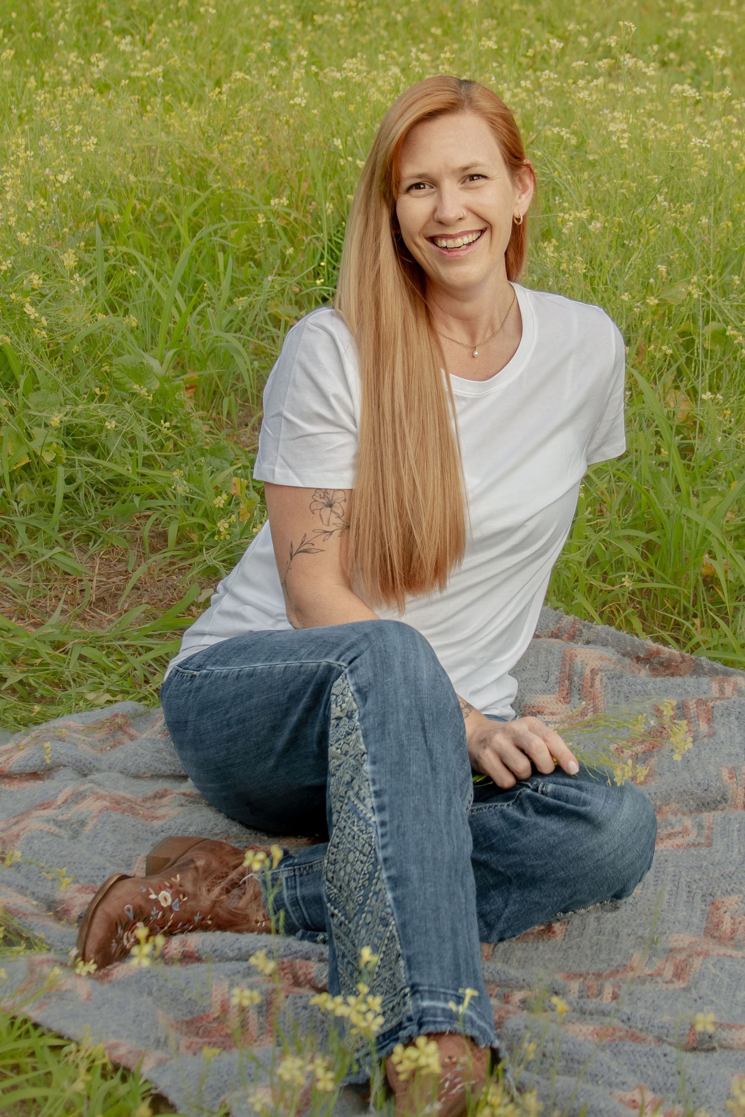 Smiling redhead woman in white tee and jeans sitting cross-legged on blanket in meadow.