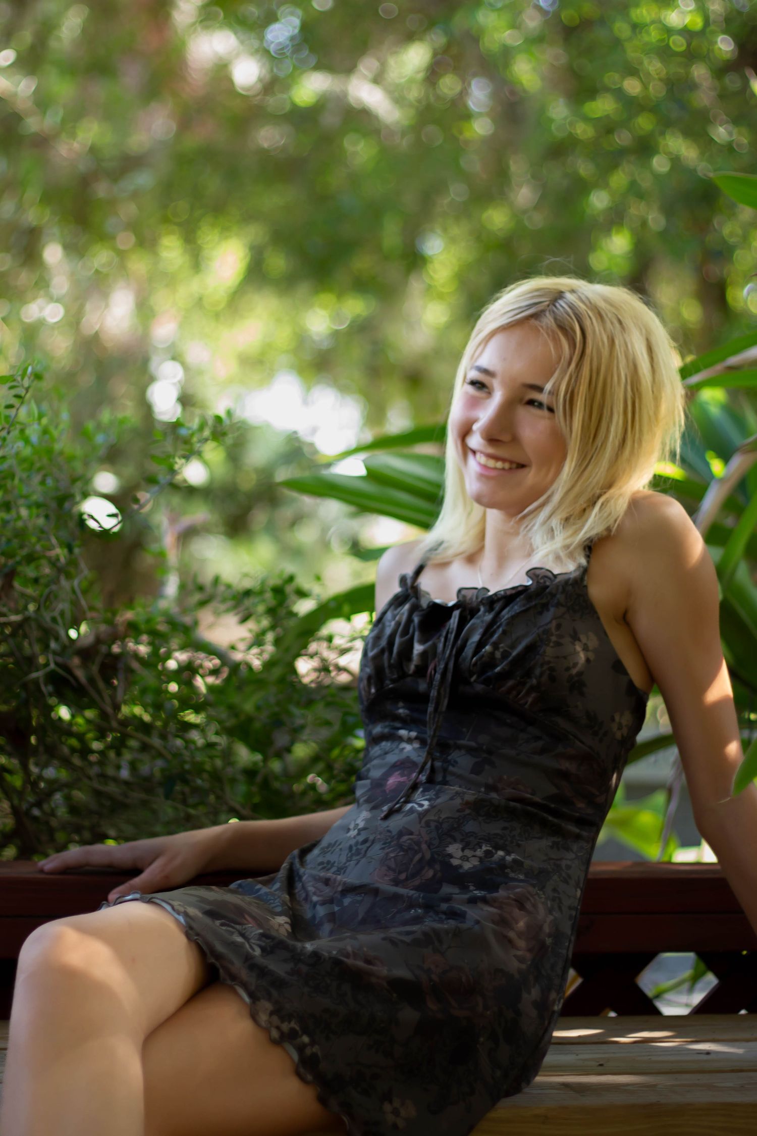 Smiling blonde woman in dark floral dress sitting on bench surrounded by lush green foliage.