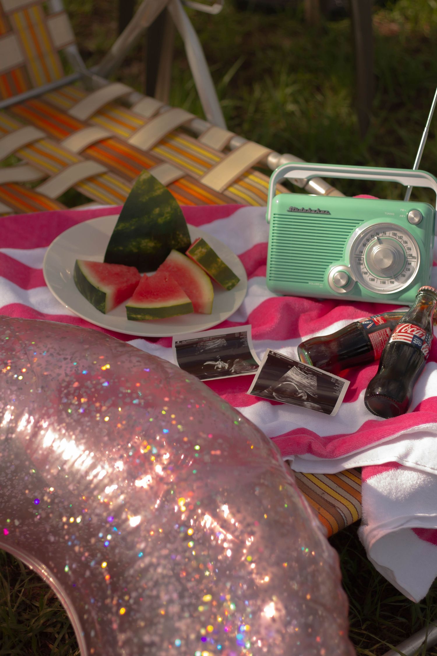 A vintage outdoor scene with watermelon slices, Coca-Cola bottles, ultrasound photos, and a retro teal radio on a vintage lawn chair.